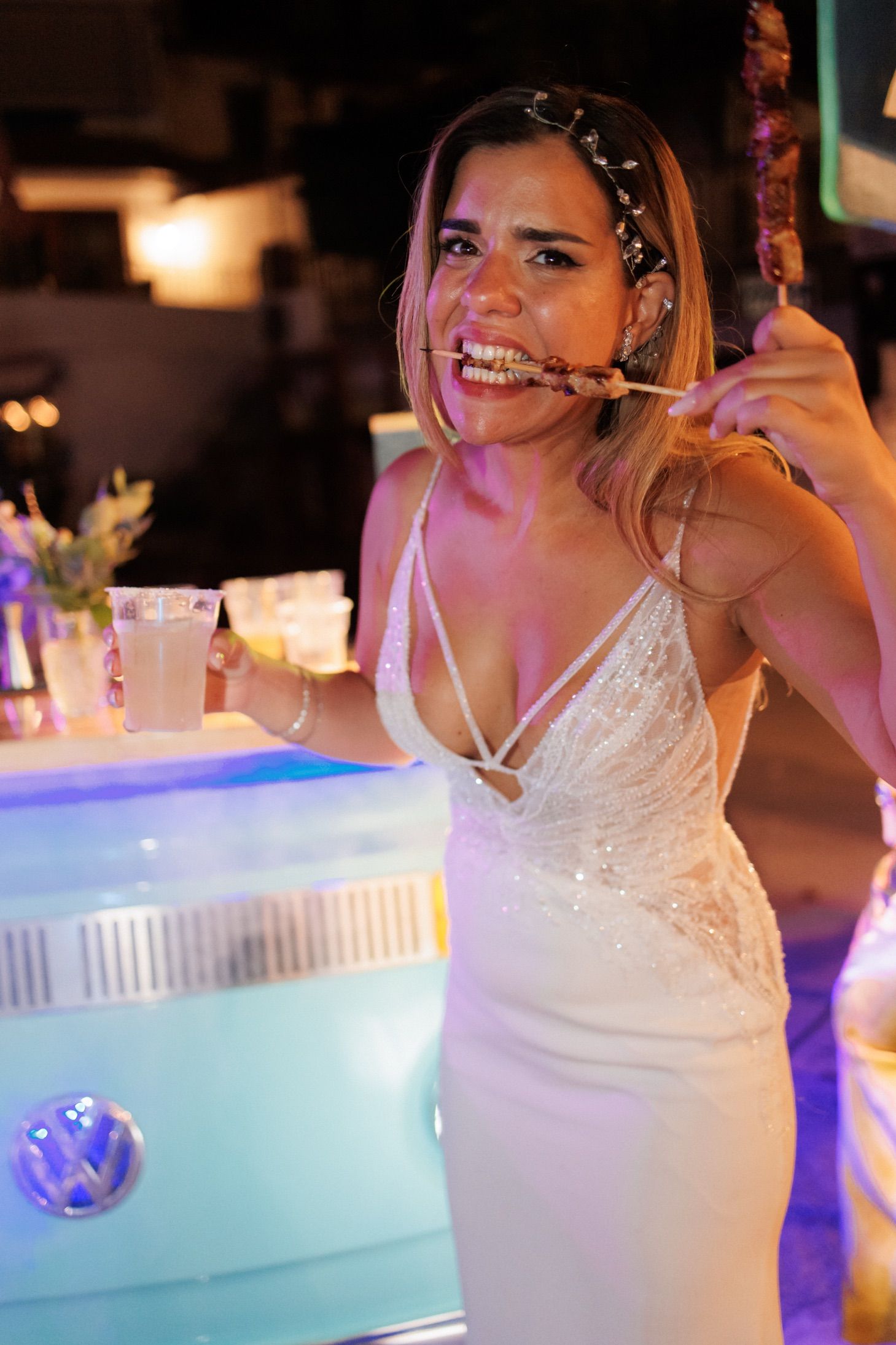 Bride in white wedding dress eating skewer food at outdoor reception bar.