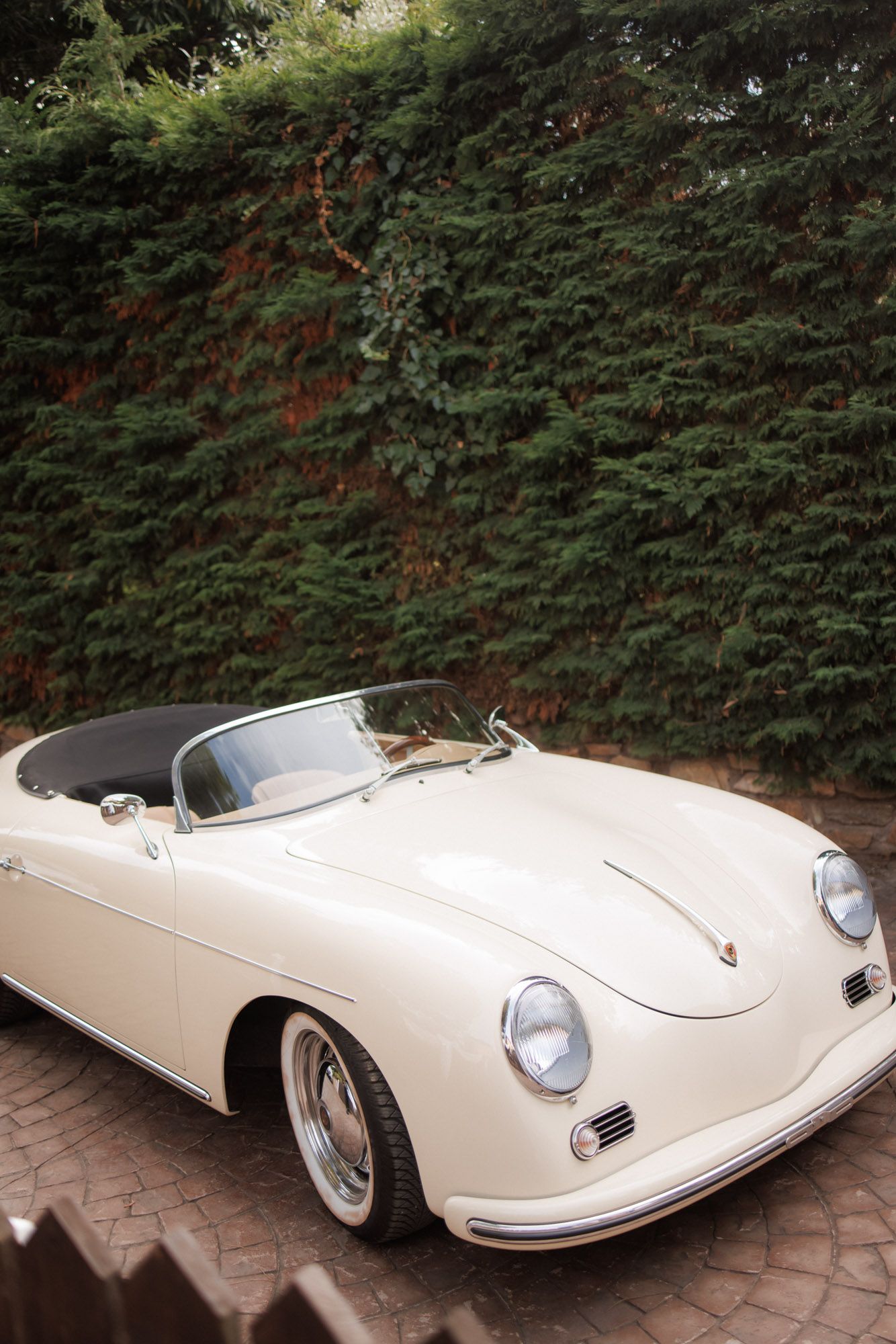Classic cream vintage Porsche 356 Speedster convertible parked against a lush green hedge backdrop.