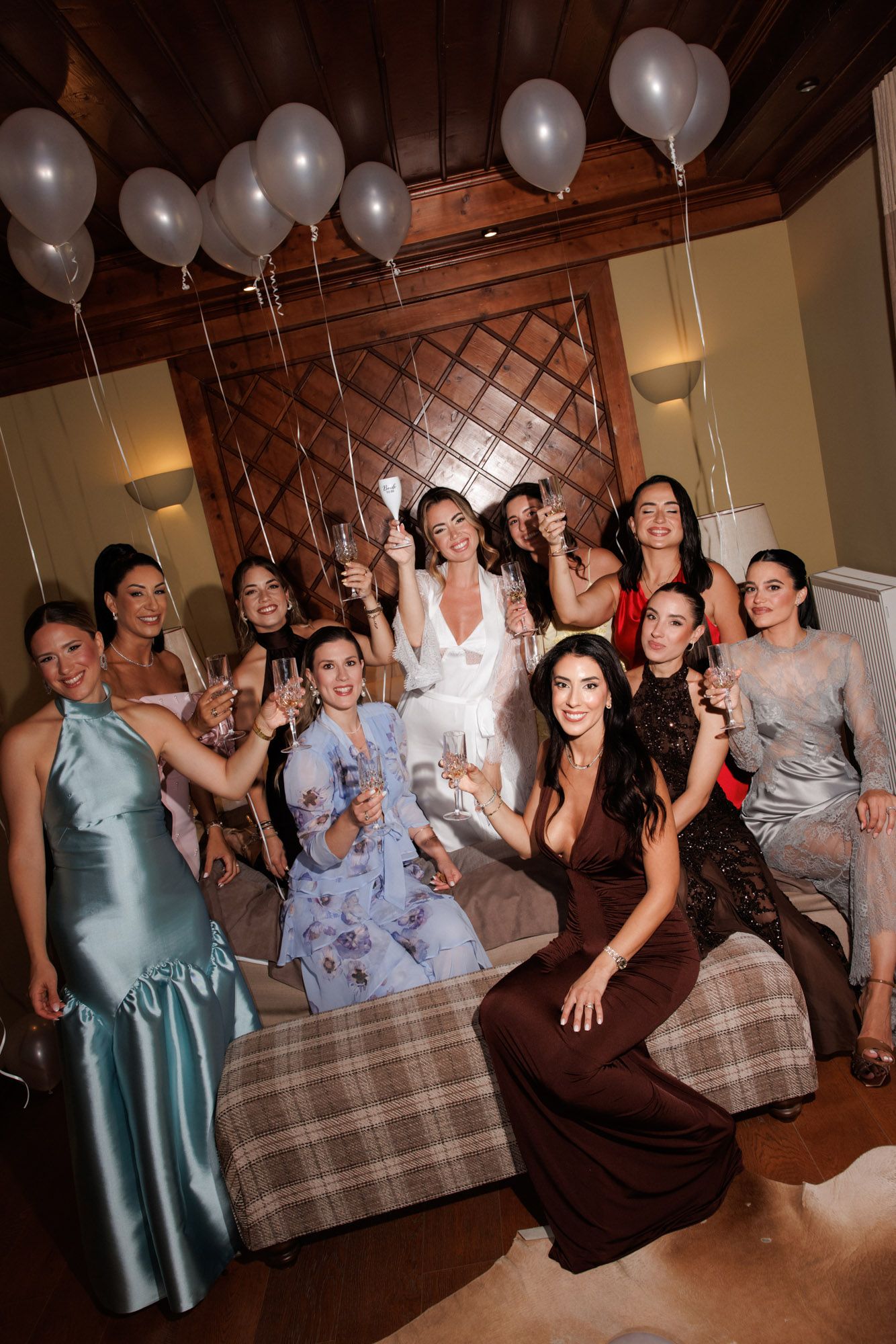 Group of elegantly dressed women celebrating at a bachelorette party with champagne and white balloons.