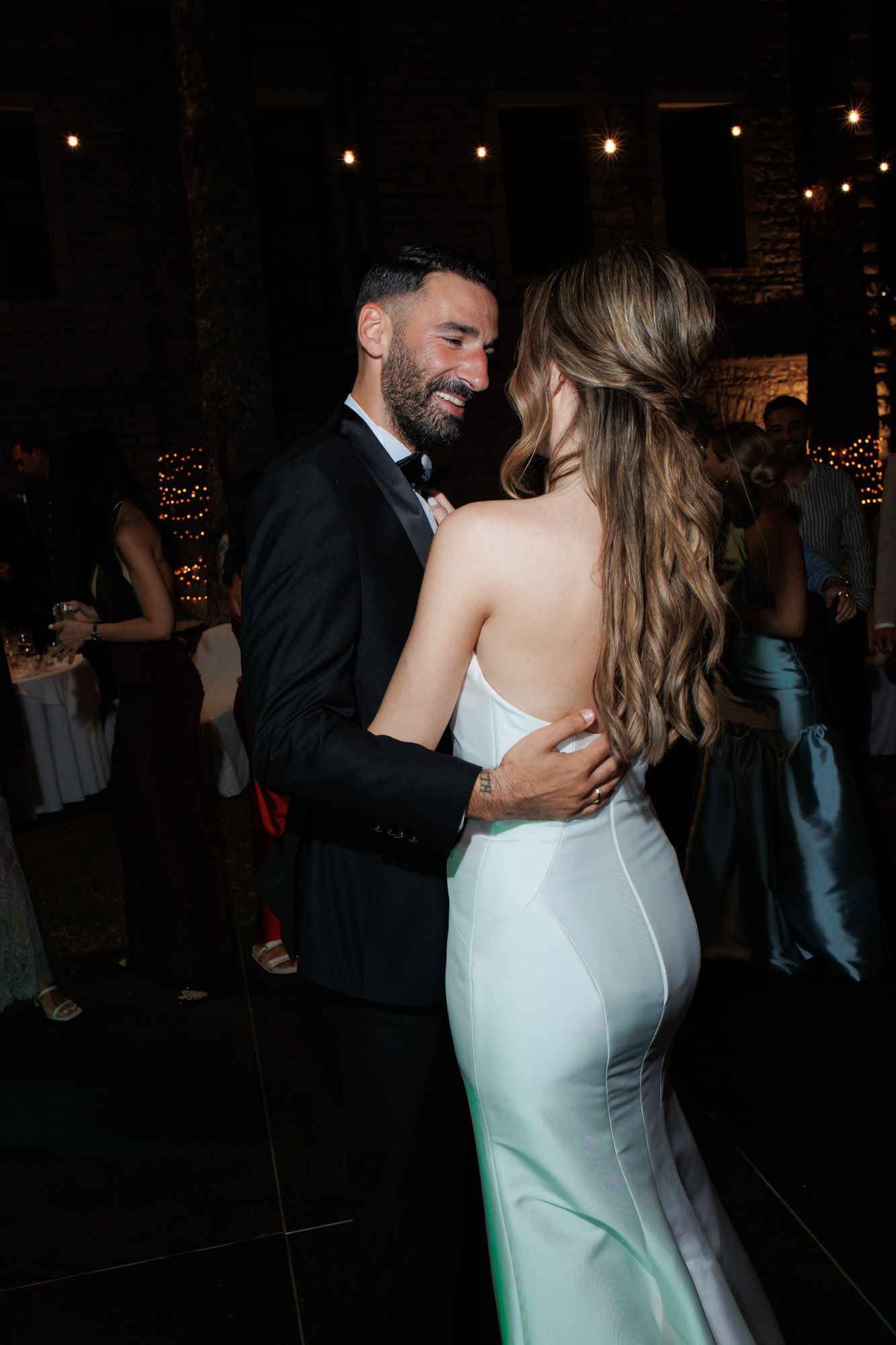 Groom in tuxedo smiling at bride in elegant backless white gown during first dance at wedding.