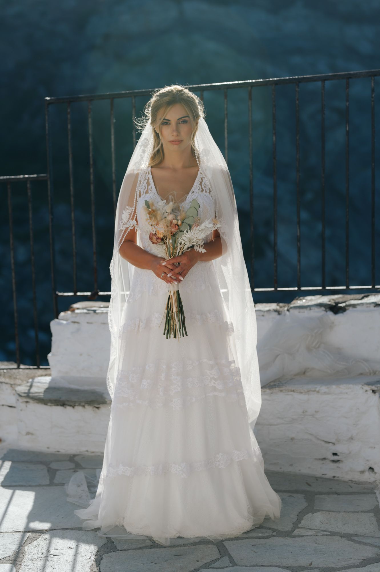 Beautiful bride in lace wedding gown and veil holds a wildflower bouquet at a sunlit outdoor venue.