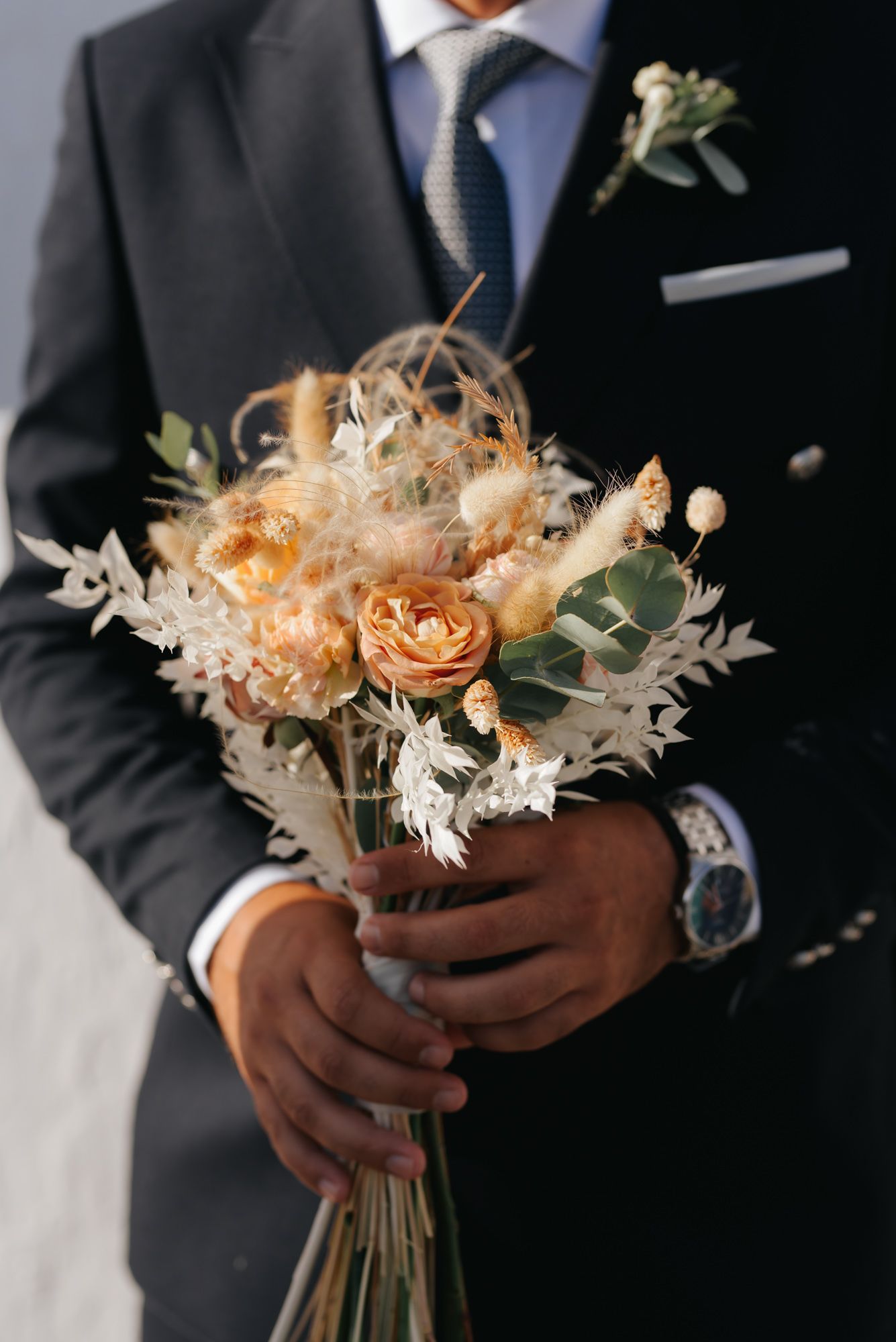 Groom in dark suit holding elegant boho wedding bouquet with dried flowers and roses.