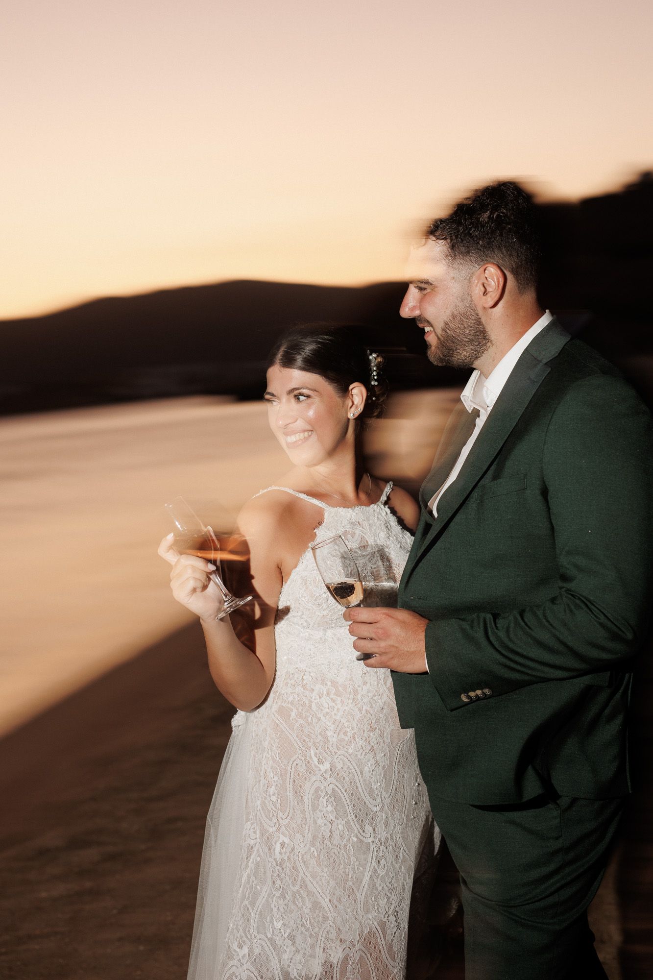 Newlywed couple in wedding attire shares drinks at golden sunset on sandy beach.