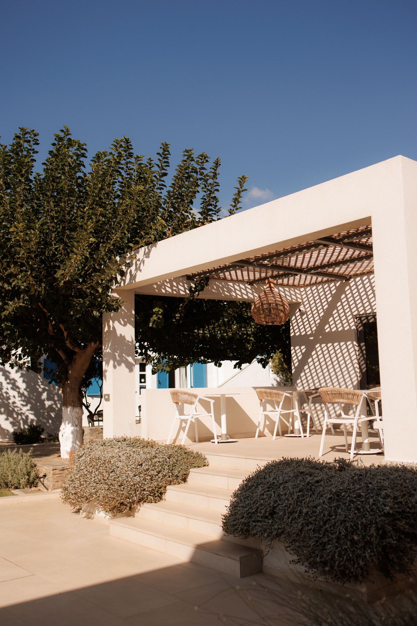 White Mediterranean villa exterior with wooden pergola, white chairs, and lush garden landscaping.