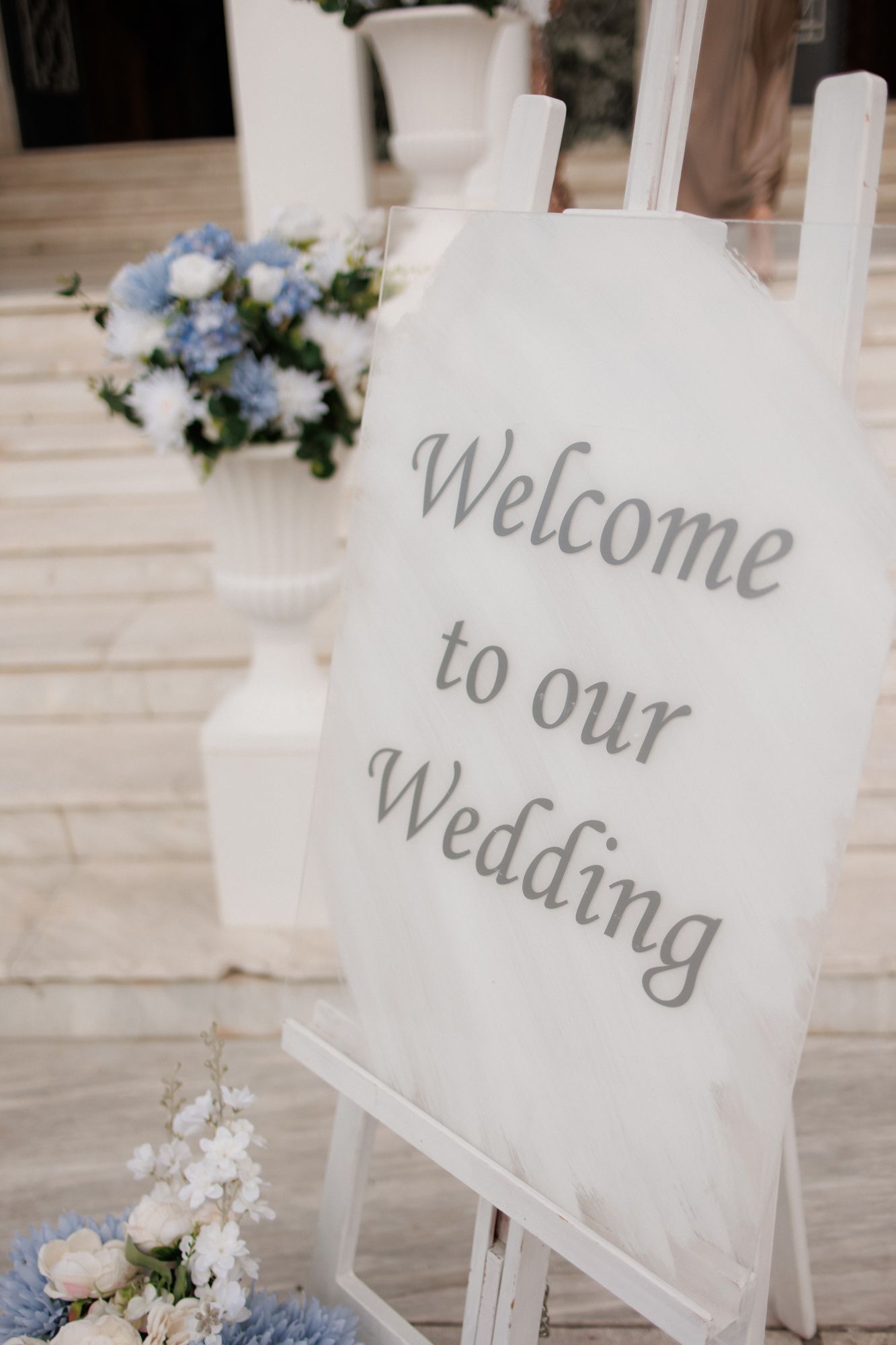Elegant white wedding welcome sign on easel with blue and white floral arrangements at outdoor ceremony.