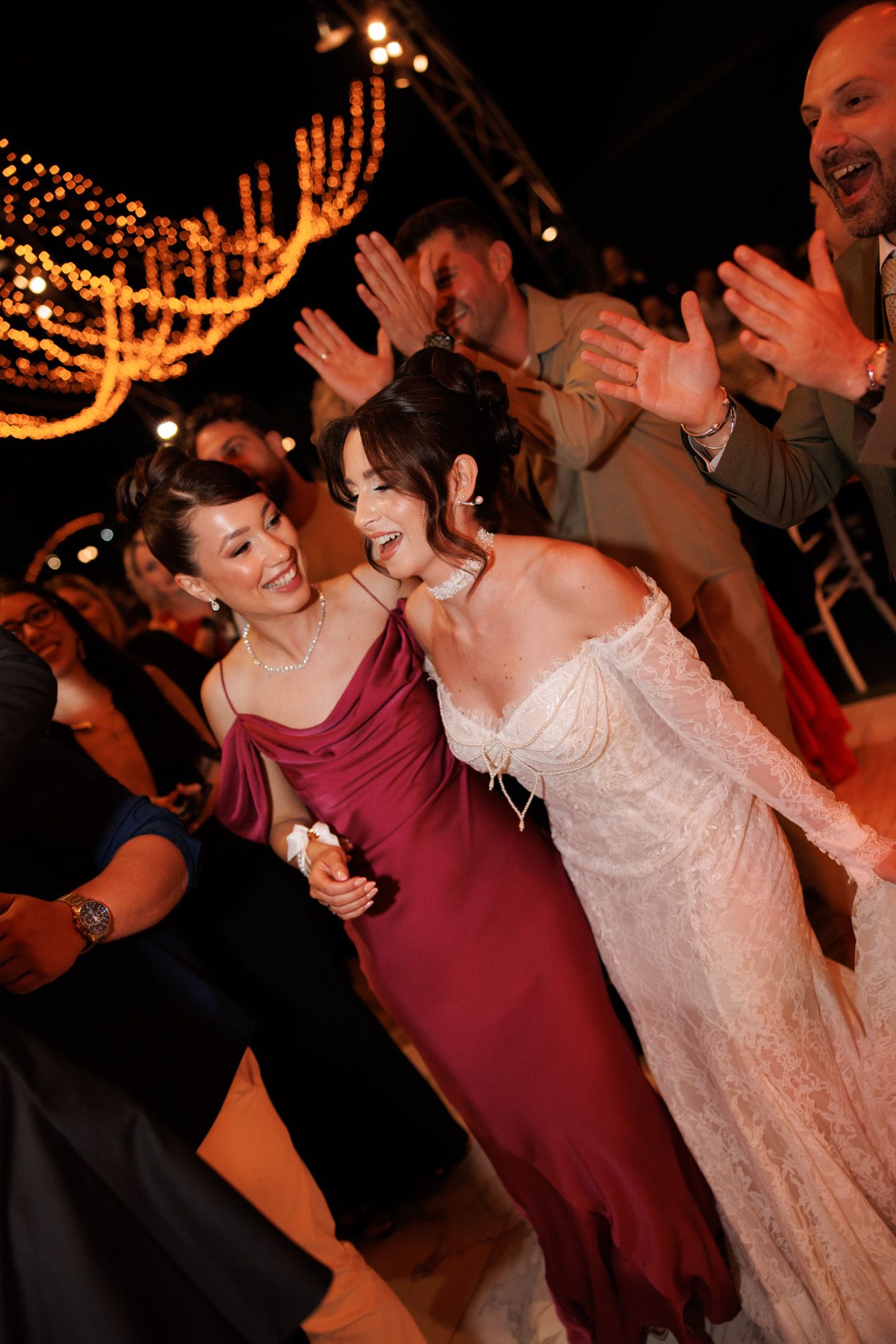 Bride in white lace gown laughing with bridesmaid in burgundy dress on wedding reception dance floor at night.