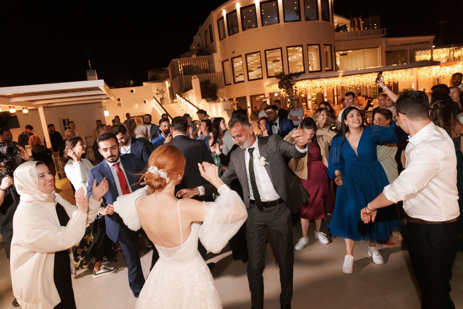 Bride and groom dance energetically with wedding guests at lively outdoor evening reception party.