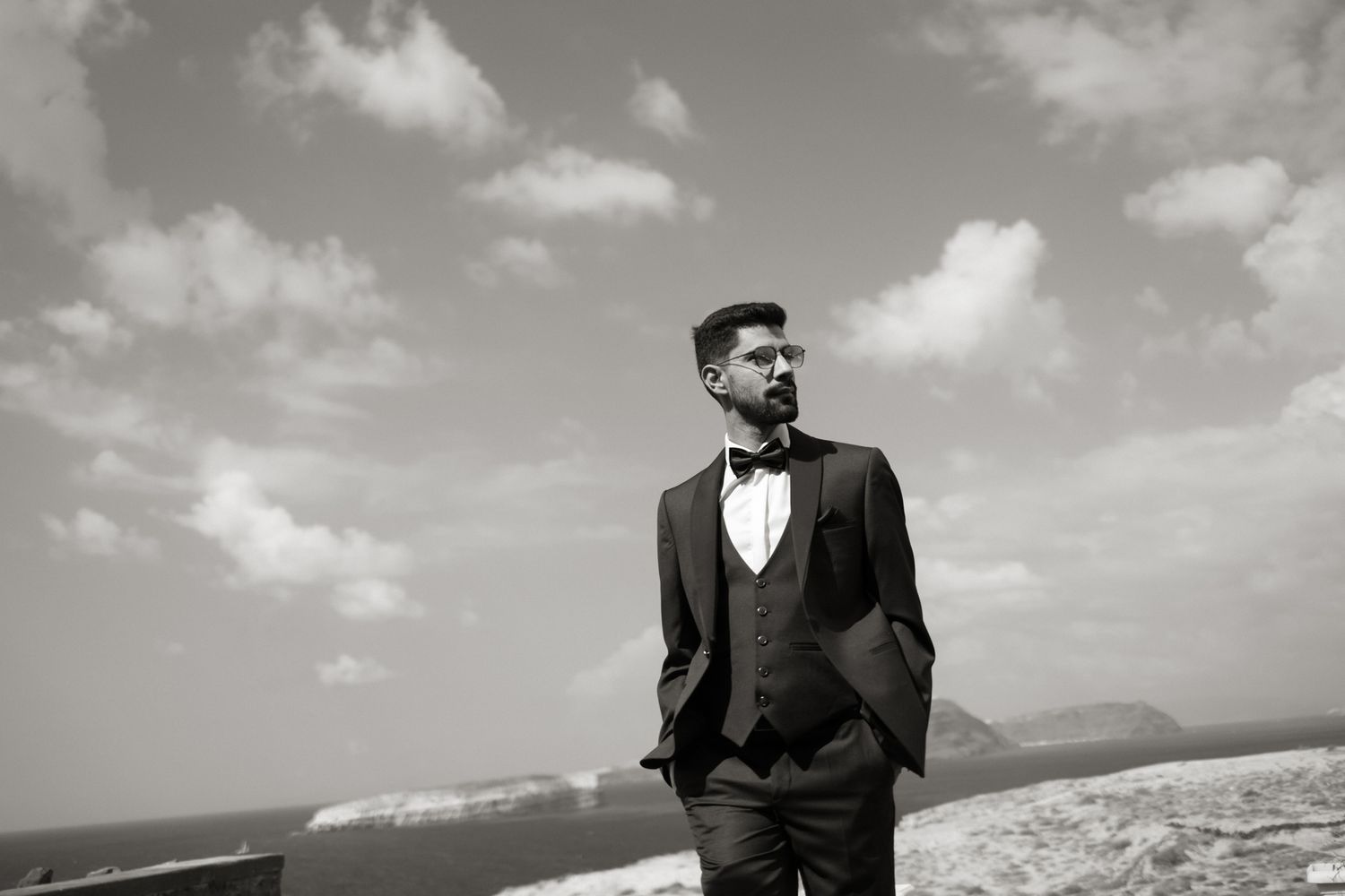 Elegant groom in black three-piece suit with bow tie posing under dramatic cloudy sky.