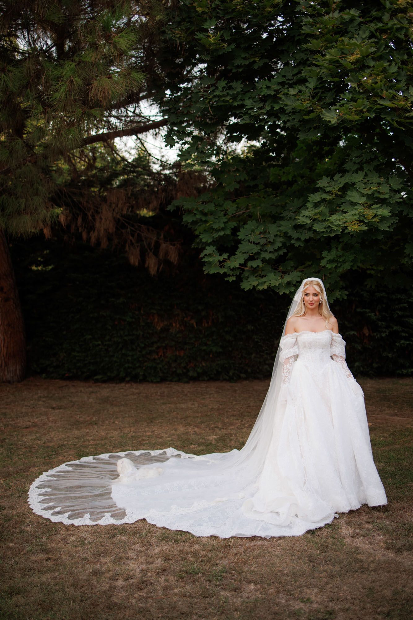 Elegant blonde bride in off-shoulder lace ball gown with long cathedral veil poses in a lush garden setting.