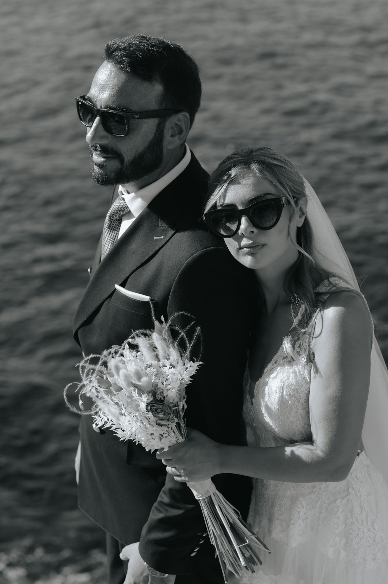 Black and white portrait of stylish bride and groom wearing sunglasses near a shimmering water backdrop.
