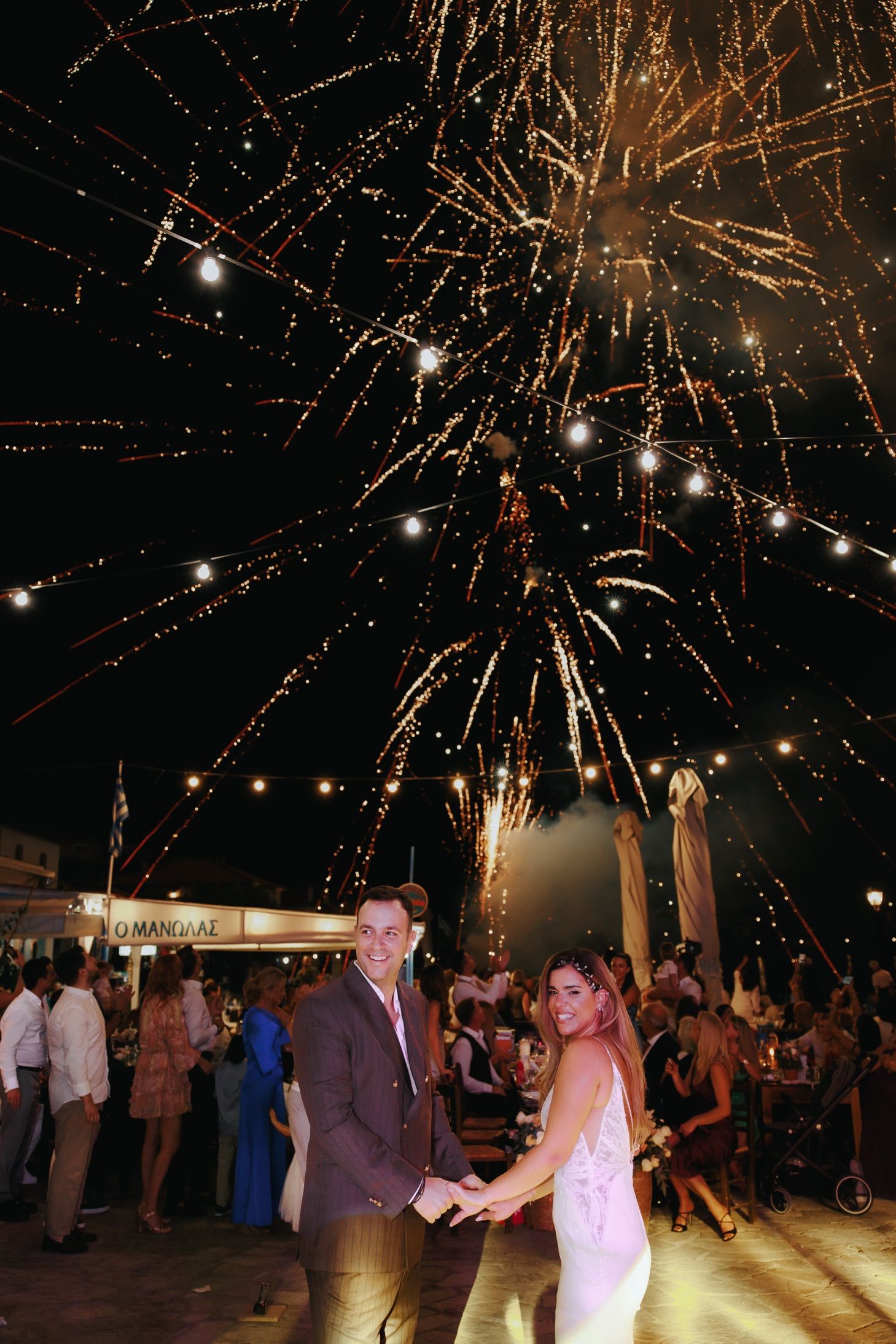 Couple holds hands smiling as golden fireworks burst overhead at elegant outdoor wedding reception.
