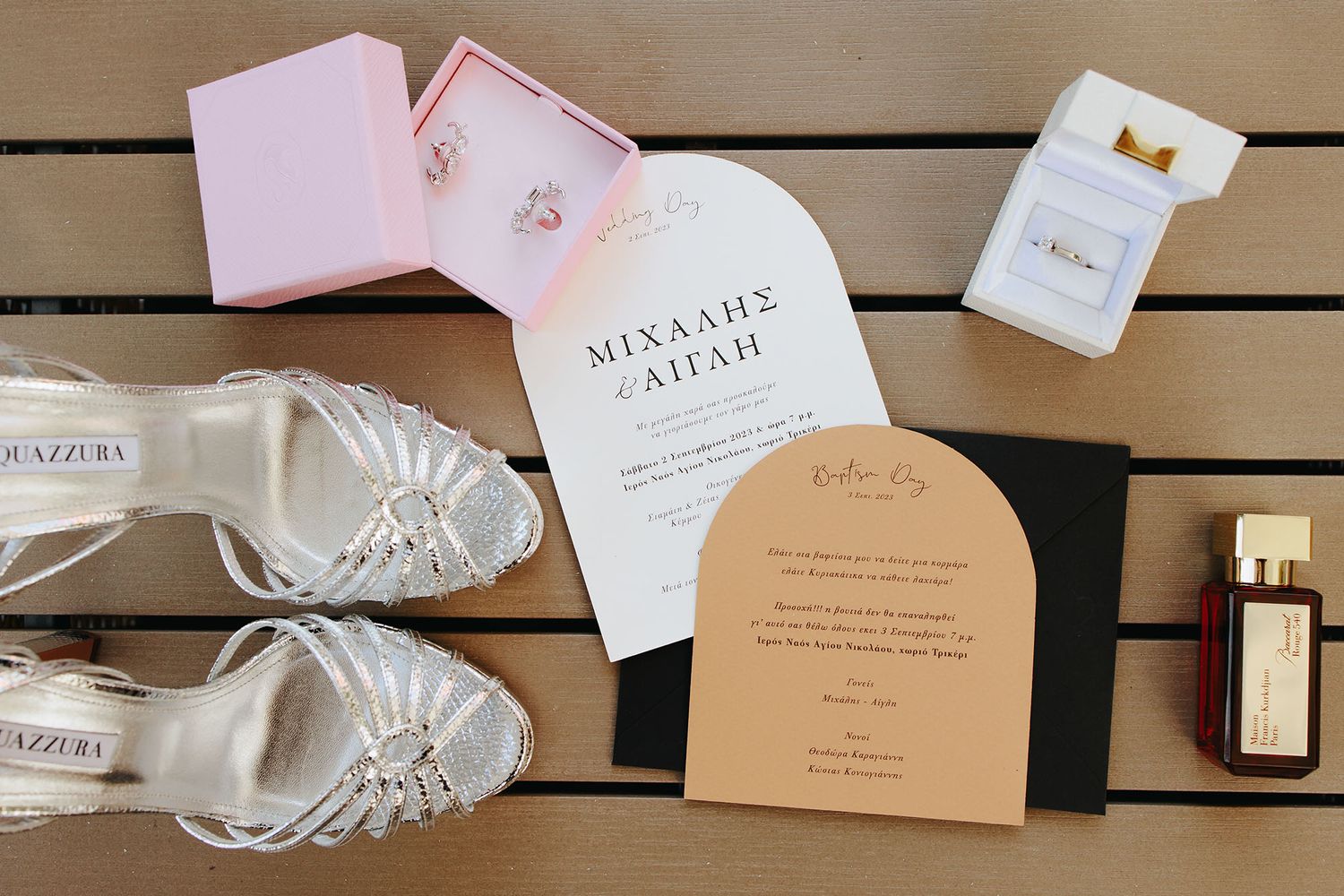 Wedding details flatlay with bridal shoes, invitations, perfume bottle and pink accessories on wooden backdrop.