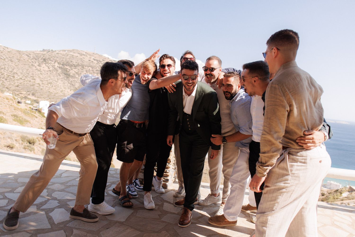 Group of groomsmen celebrating together outdoors with Mediterranean sea view in background.
