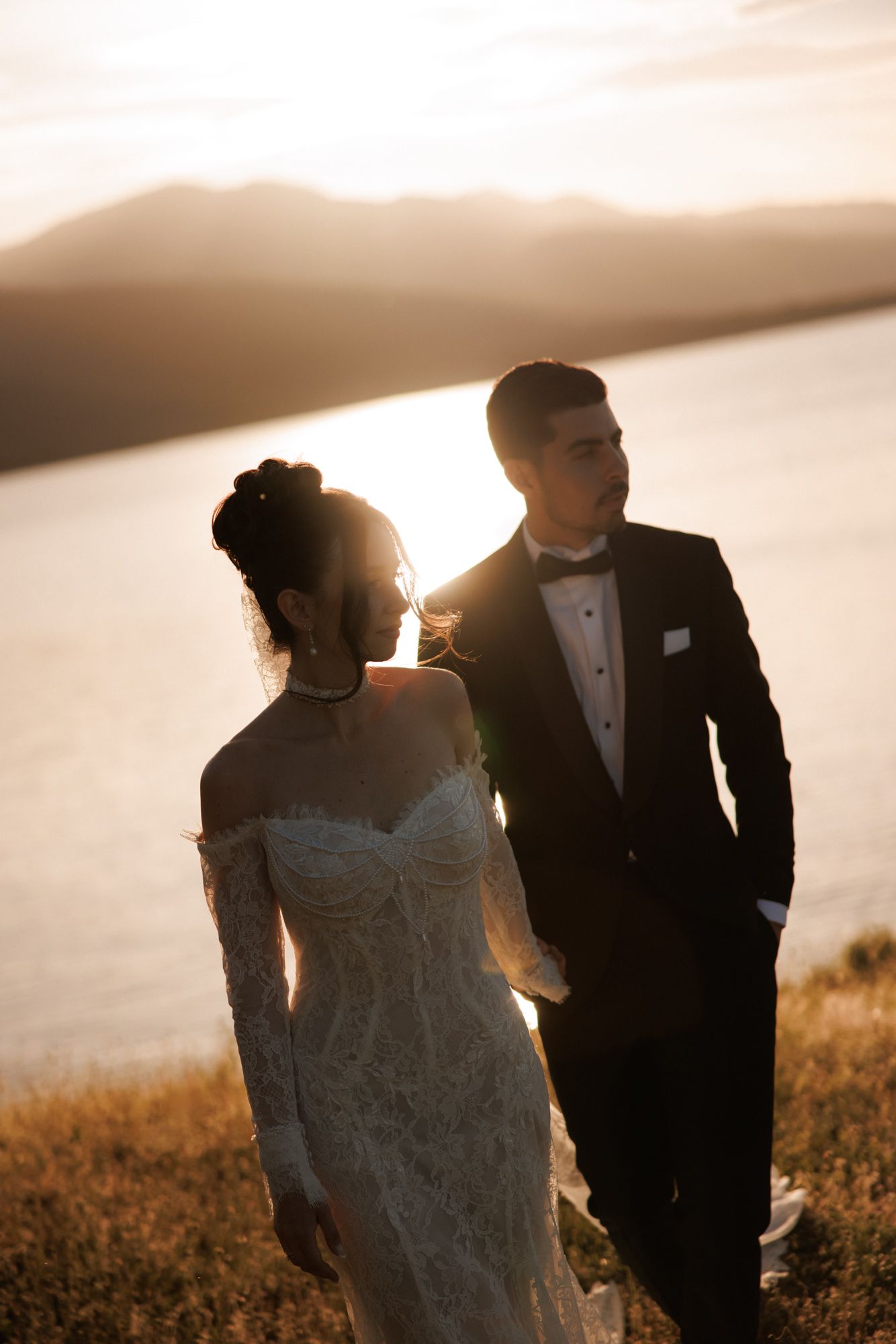 Bride in lace off-shoulder gown and groom in tuxedo stand together at golden sunset by a lake.