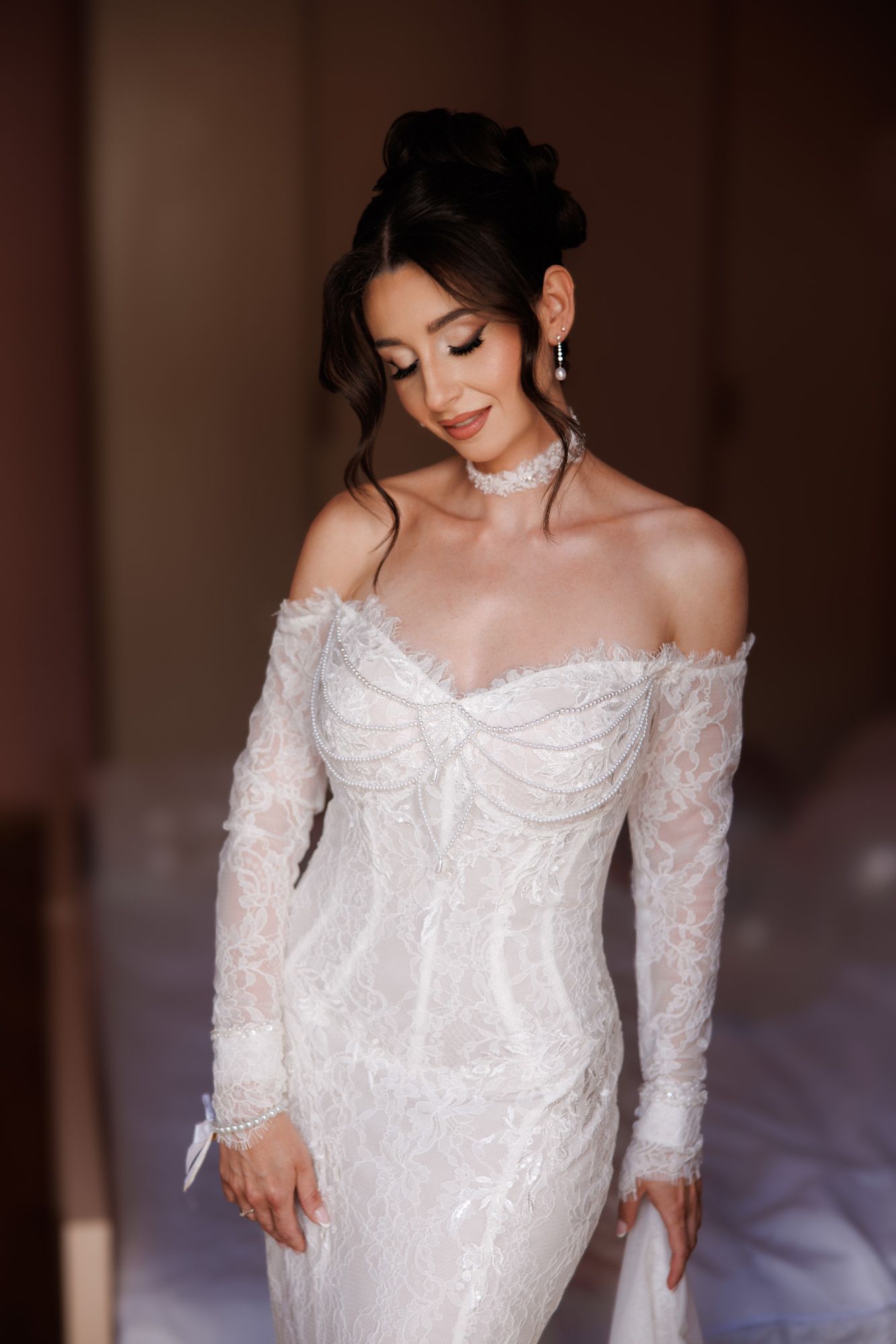 Elegant bride in off-shoulder white lace wedding gown with long sleeves, updo hairstyle, and delicate jewelry.