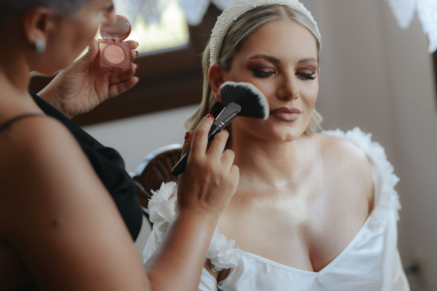 Makeup artist applying blush to a bride in a white dress and headband on her wedding day.