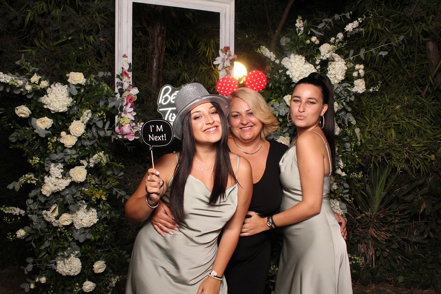 Three women pose at a floral photo booth with fun props at a wedding celebration.