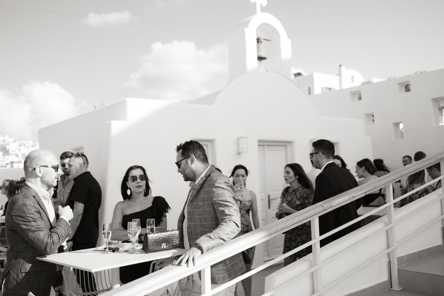 Black and white photo of wedding guests socializing near white Greek chapel with bell tower.