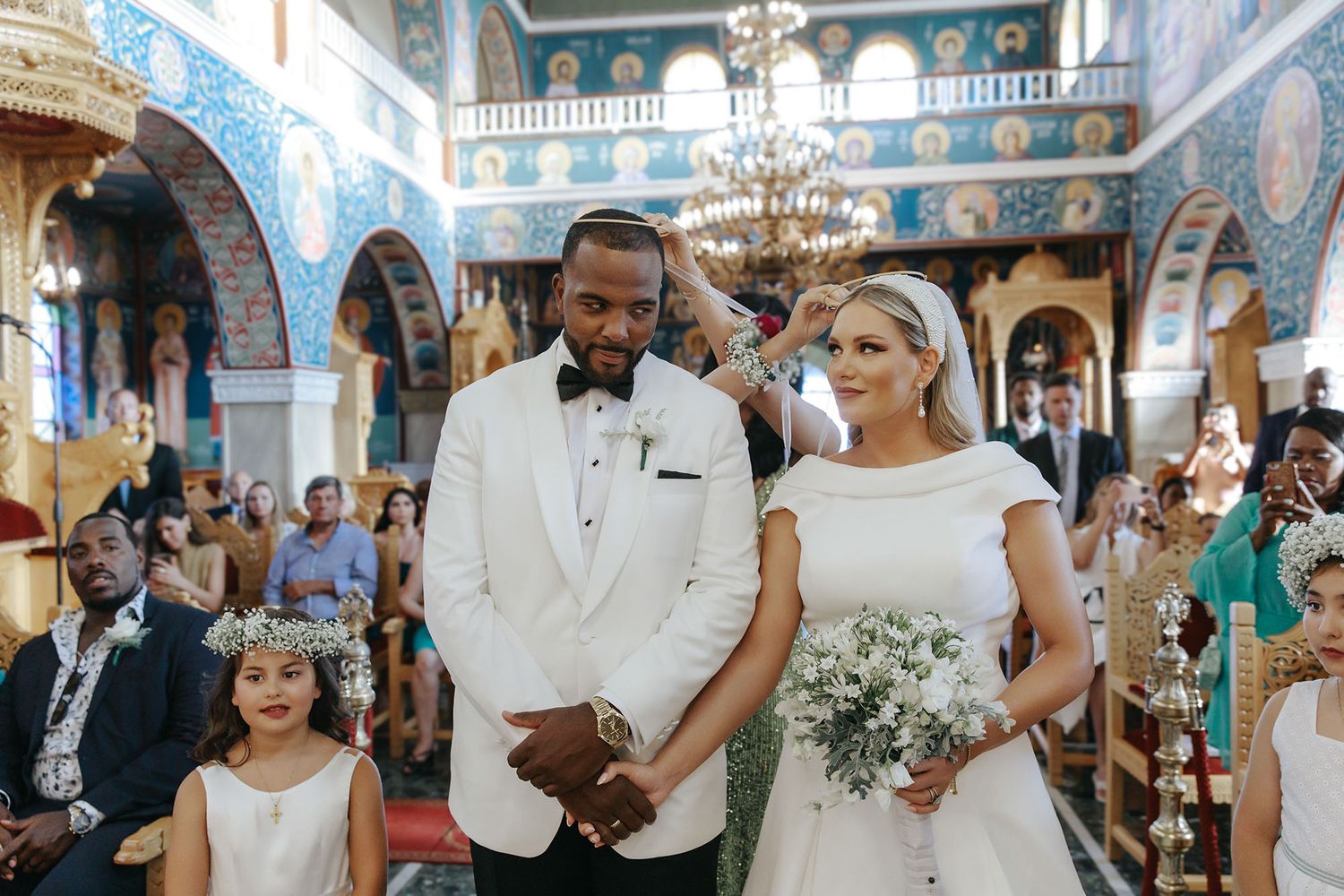 Couple celebrates joyous wedding ceremony inside ornate orthodox church with blue frescoes and golden chandelier.