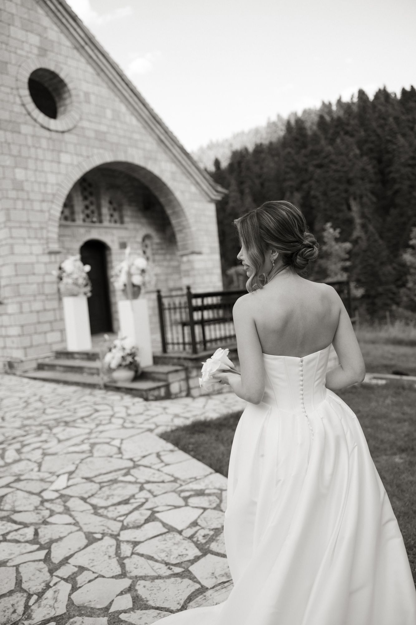 Black and white photo of a bride in a strapless gown walking toward a stone chapel.