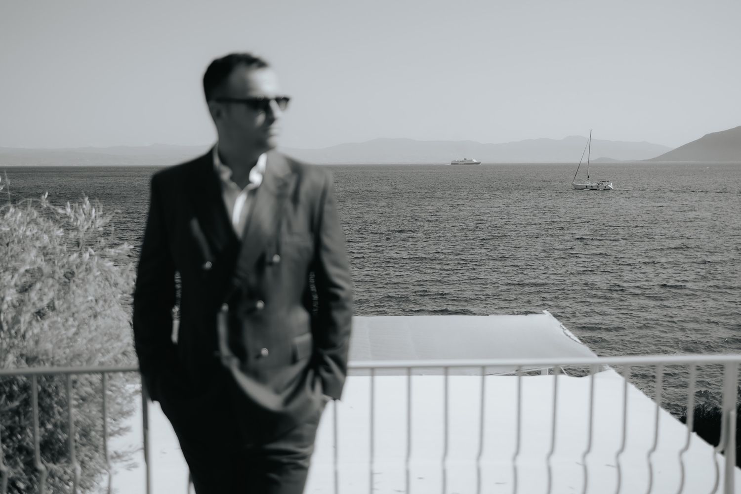 Stylish man in a dark suit and sunglasses standing on a seaside balcony in black and white photo.