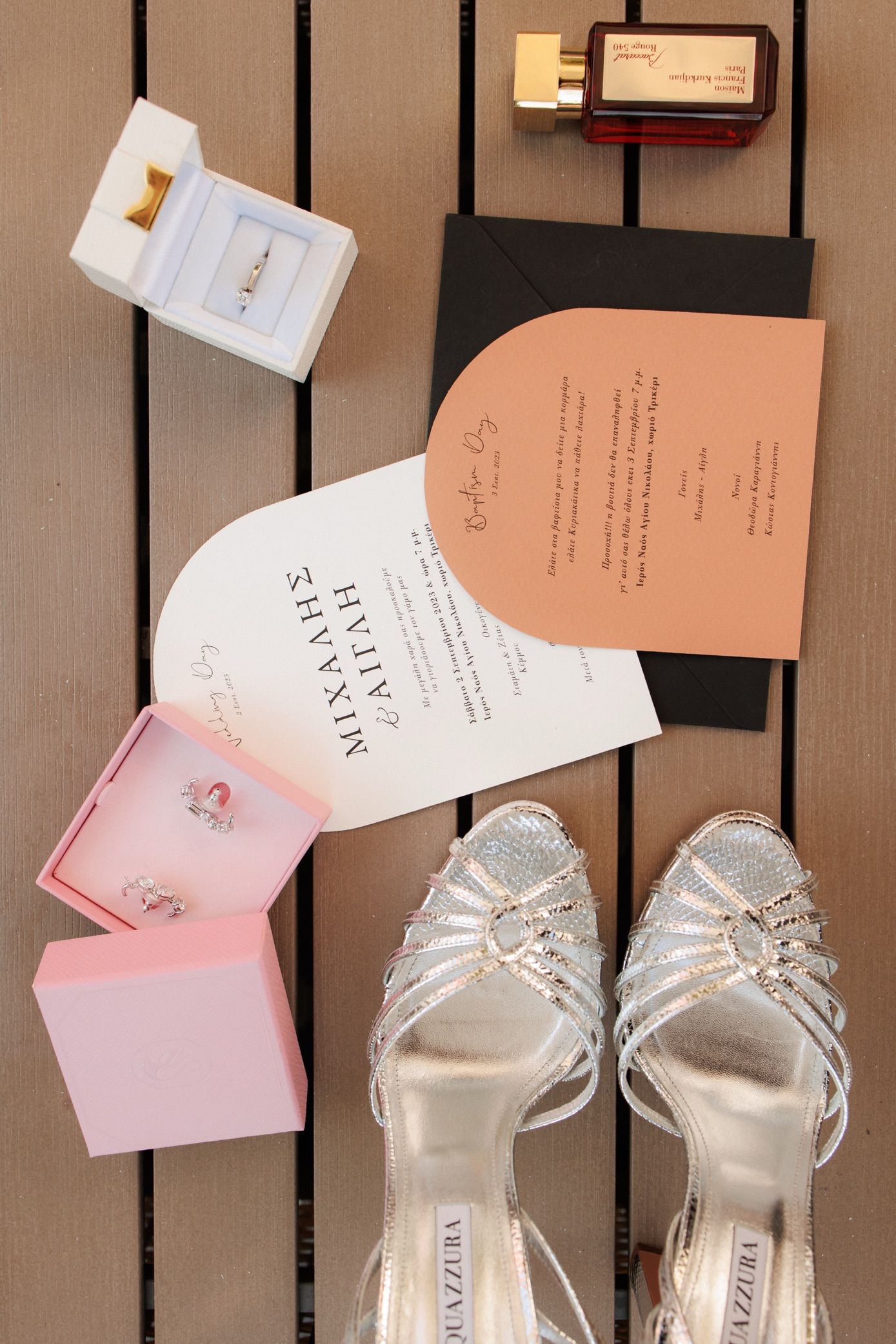 Wedding flat lay featuring silver heels, rings, invitations, and perfume on a wooden surface.