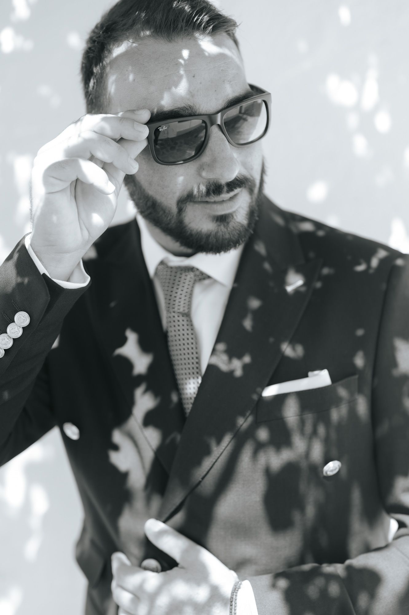 Black and white portrait of bearded groom in suit adjusting sunglasses with confetti falling.