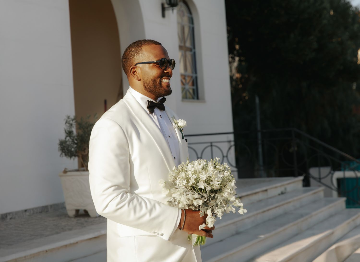 Smiling groom in white tuxedo holding white floral bouquet standing on steps outside a wedding venue.