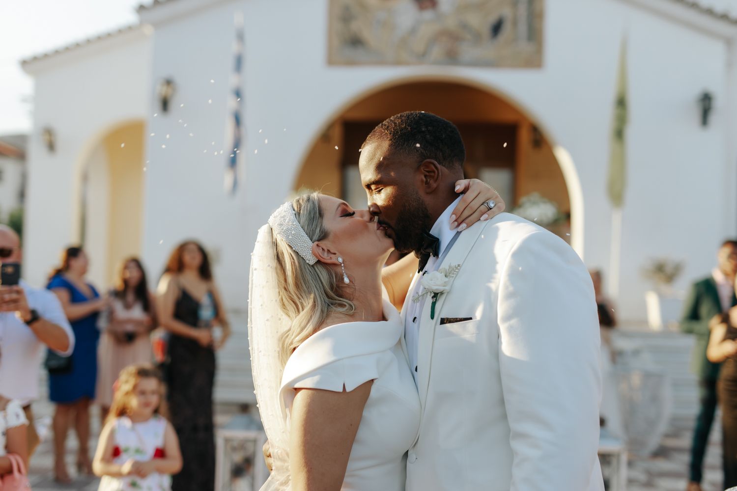 Bride and groom share a first kiss outside a white chapel as guests celebrate their wedding ceremony.