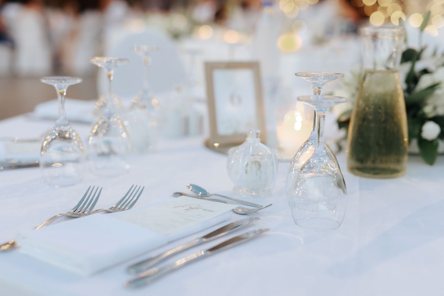 Elegant wedding table setting with white linens, crystal glasses, and silver cutlery in a softly lit reception venue.