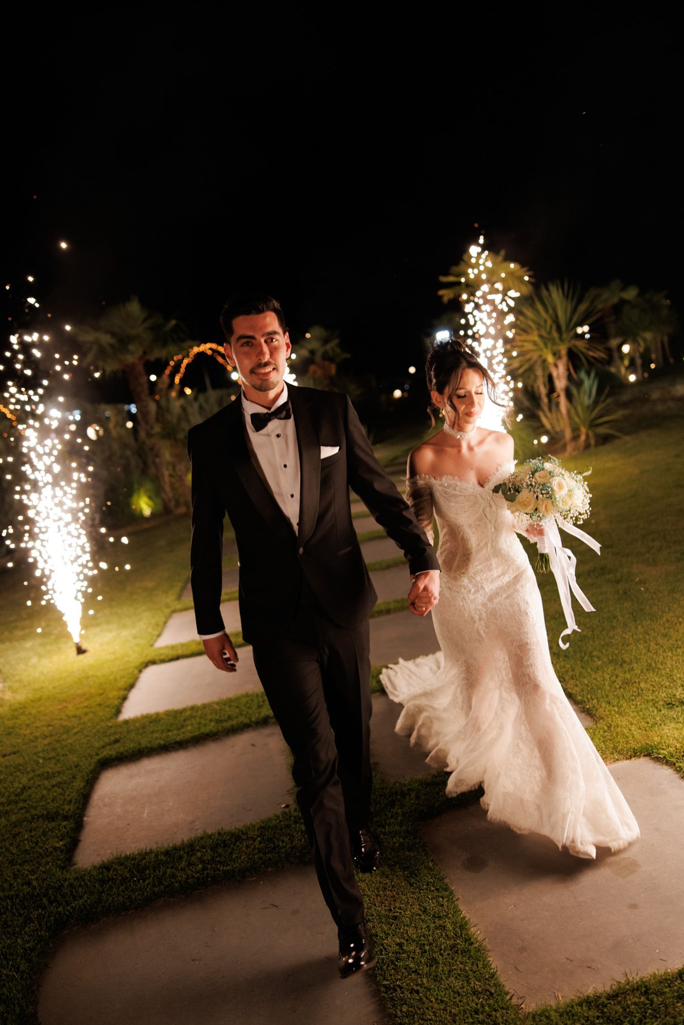 Bride and groom walk through sparkling fireworks at night wedding celebration, bride in white gown holding bouquet.