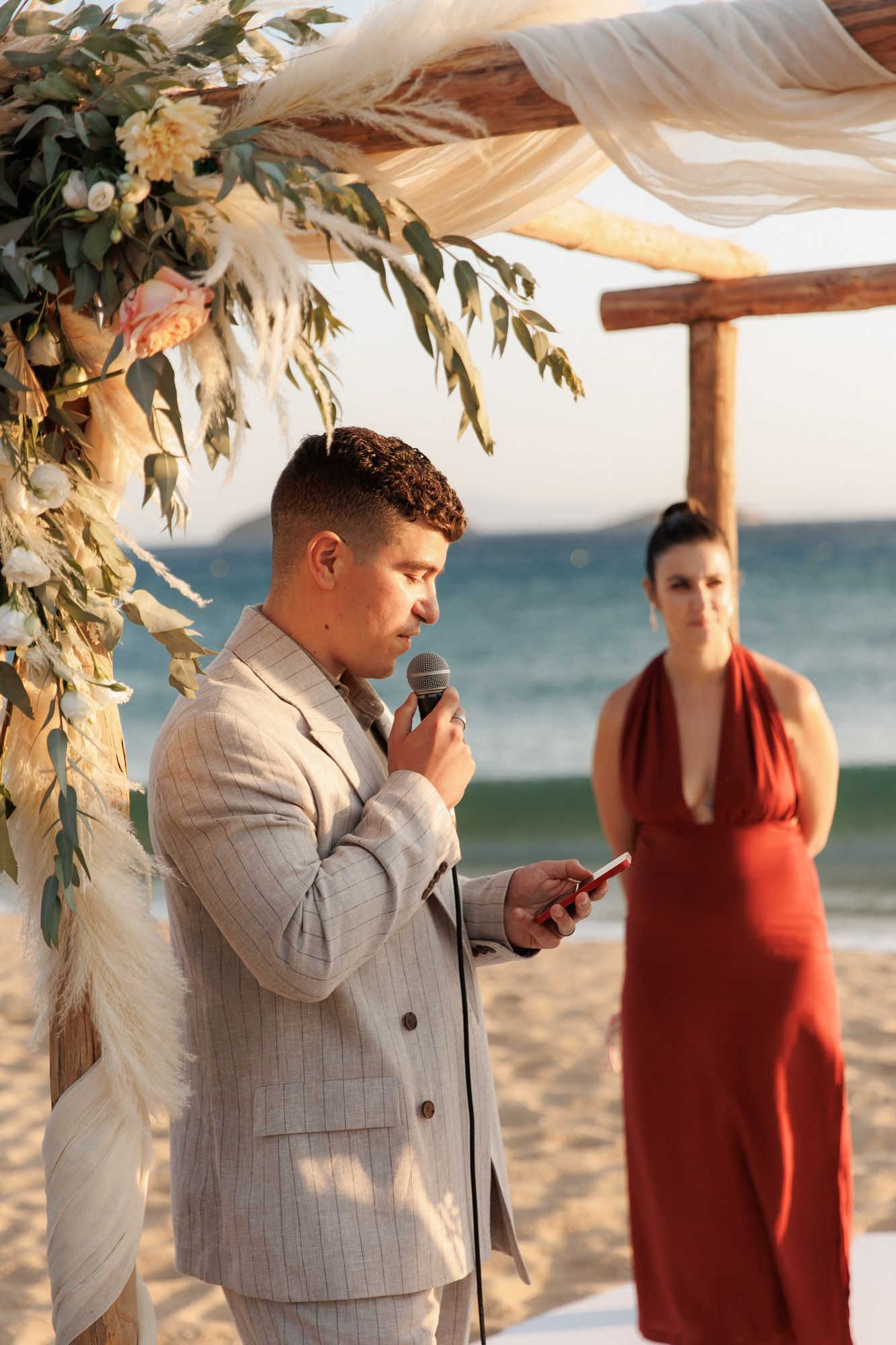 Man in gray suit speaks into microphone at beach wedding ceremony with woman in red dress behind.