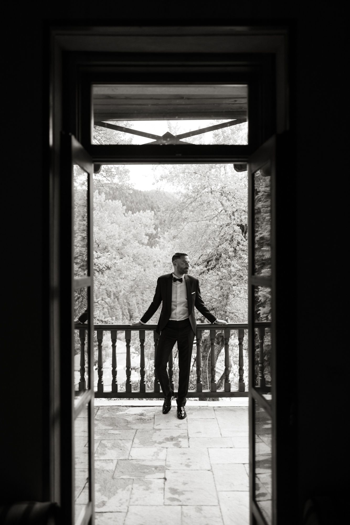 Black and white silhouette of groom in tuxedo standing on balcony with lush trees in background.