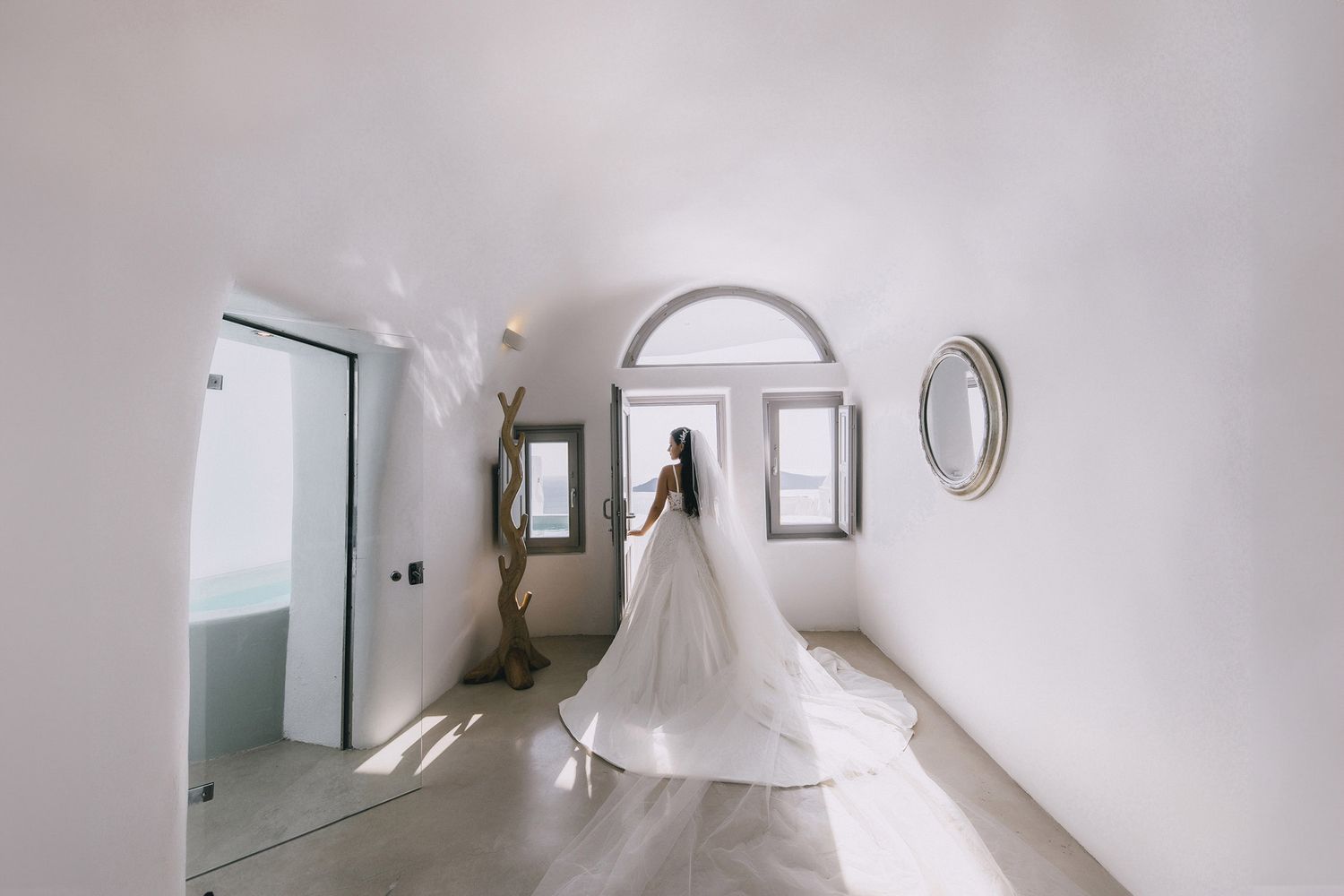 Bride in white wedding gown standing by arched window in elegant white Santorini villa room with natural light.
