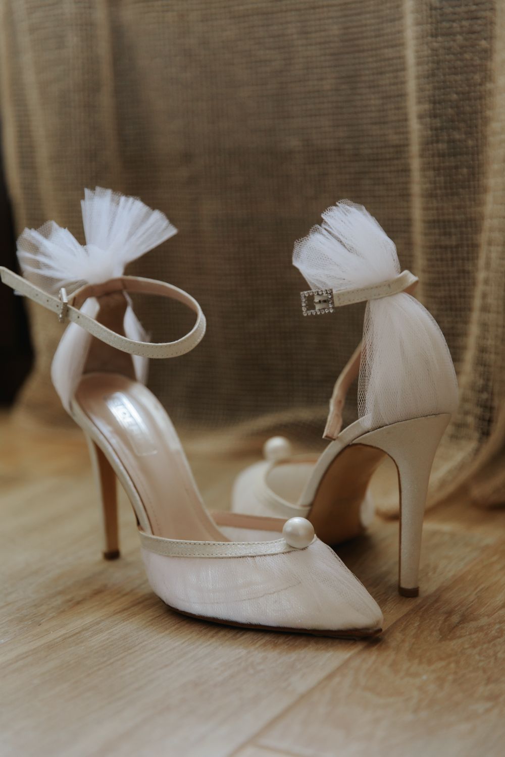 Elegant white bridal heels with tulle bow accents and pearl details on a wooden floor.