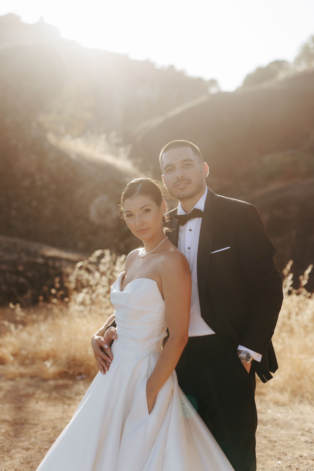 Bride in strapless white gown and groom in black tuxedo pose together at golden hour with sunlit hills behind them.
