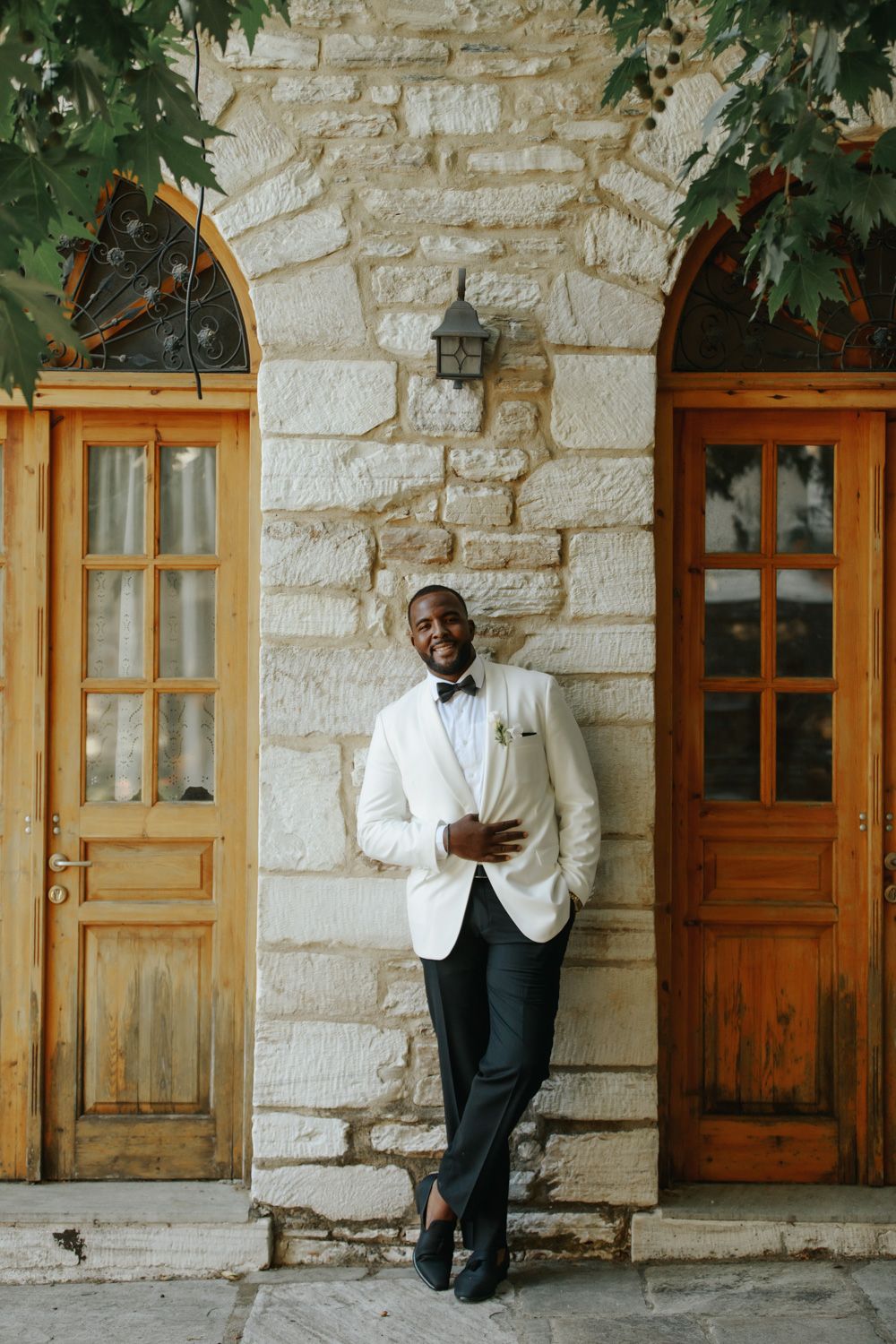 Smiling groom in white tuxedo jacket and black bow tie leaning against stone wall between wooden doors.