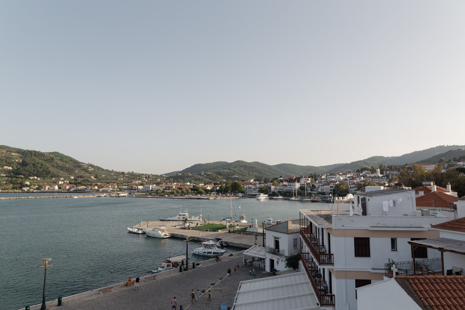 Scenic view of a Greek island harbor with white buildings, boats, and green hills at dusk.