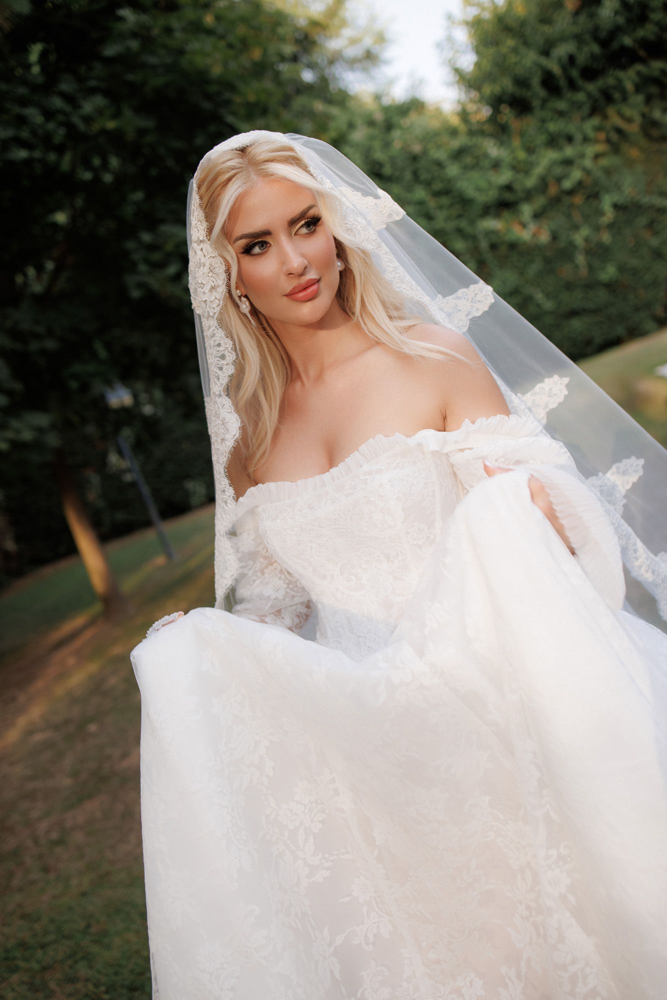 Beautiful blonde bride in off-shoulder lace wedding gown with lace-edged veil poses softly in an outdoor garden.