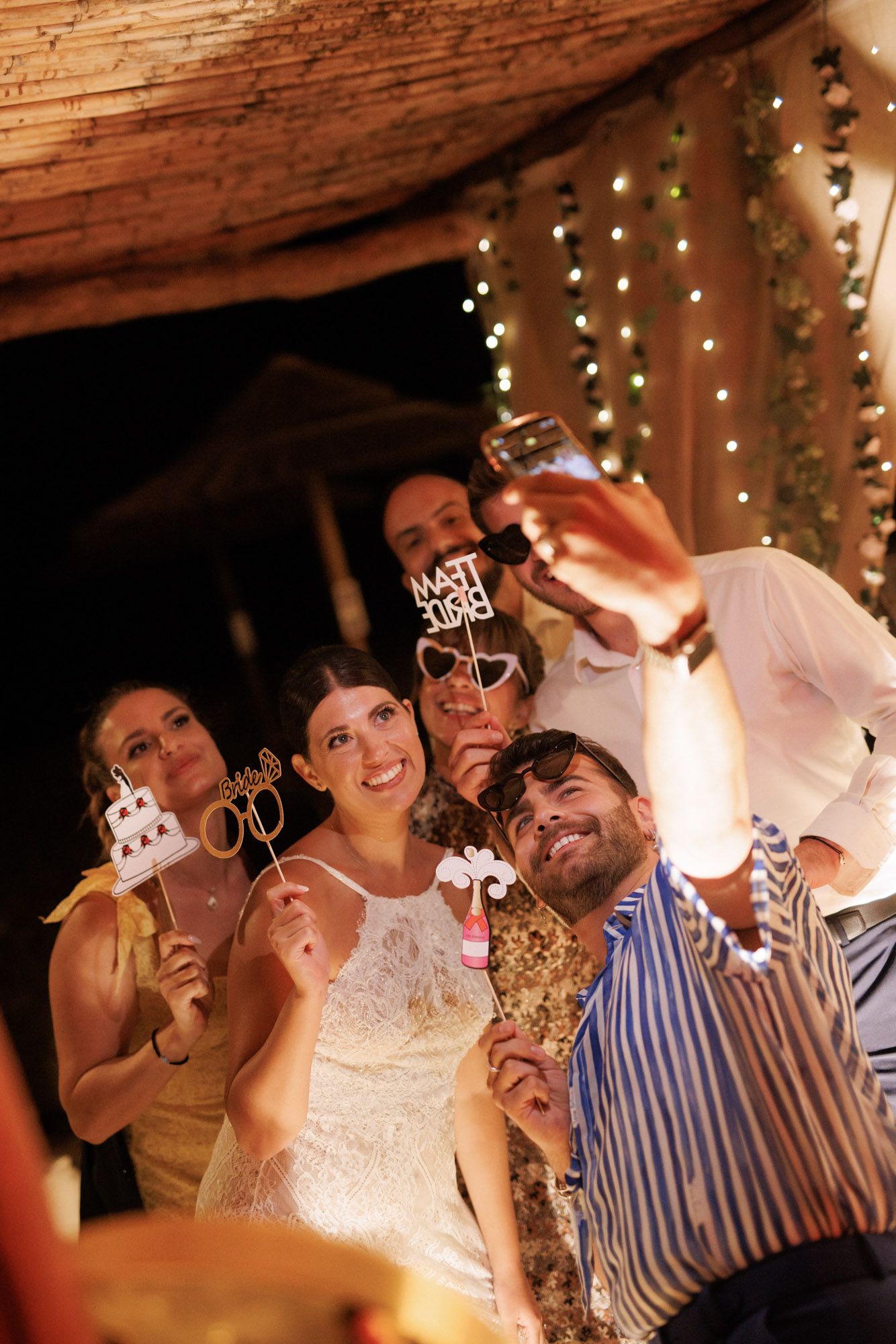 Wedding party takes fun selfie with photo booth props at beach reception celebration.