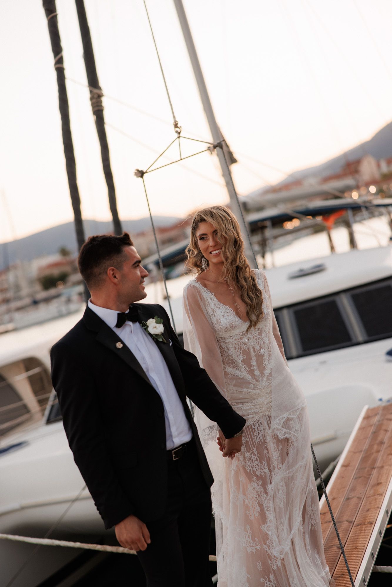 Elegant wedding couple holds hands on a marina dock surrounded by sailboats at golden hour.