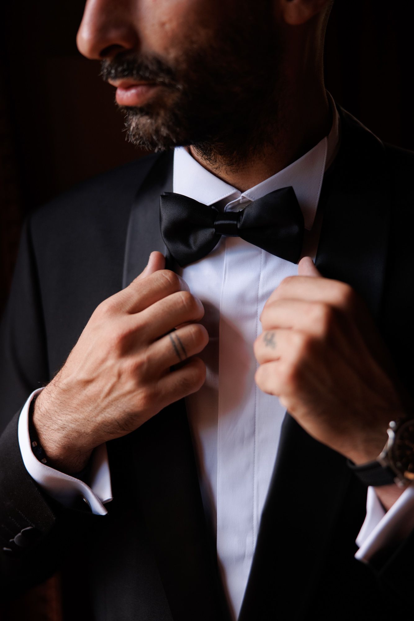 Close-up of bearded groom in black tuxedo adjusting his black bow tie with both hands.