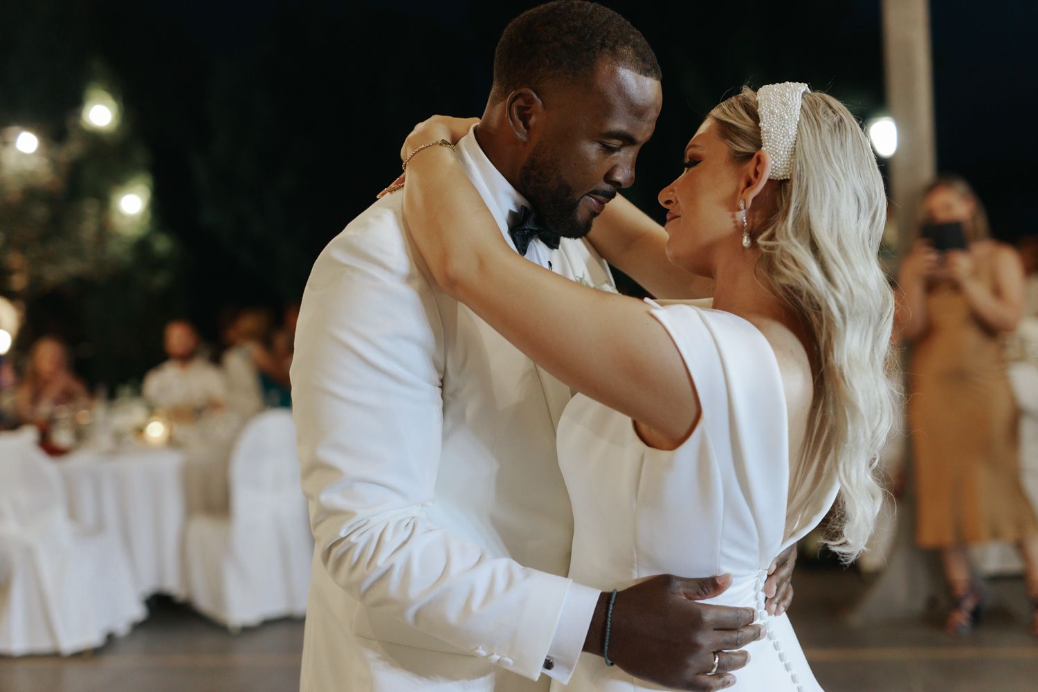 Couple sharing first dance at wedding reception, groom in white suit, bride in elegant white gown, romantic outdoor setting.