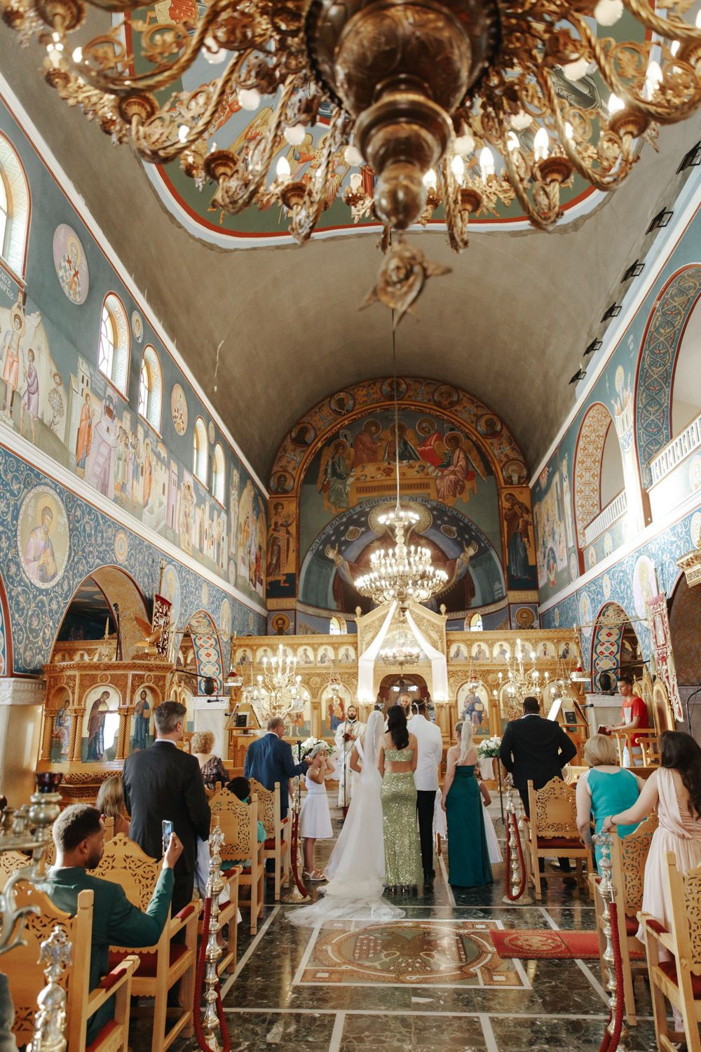 Bride walking down the aisle in an ornate Orthodox church with golden chandeliers and colorful frescoes.