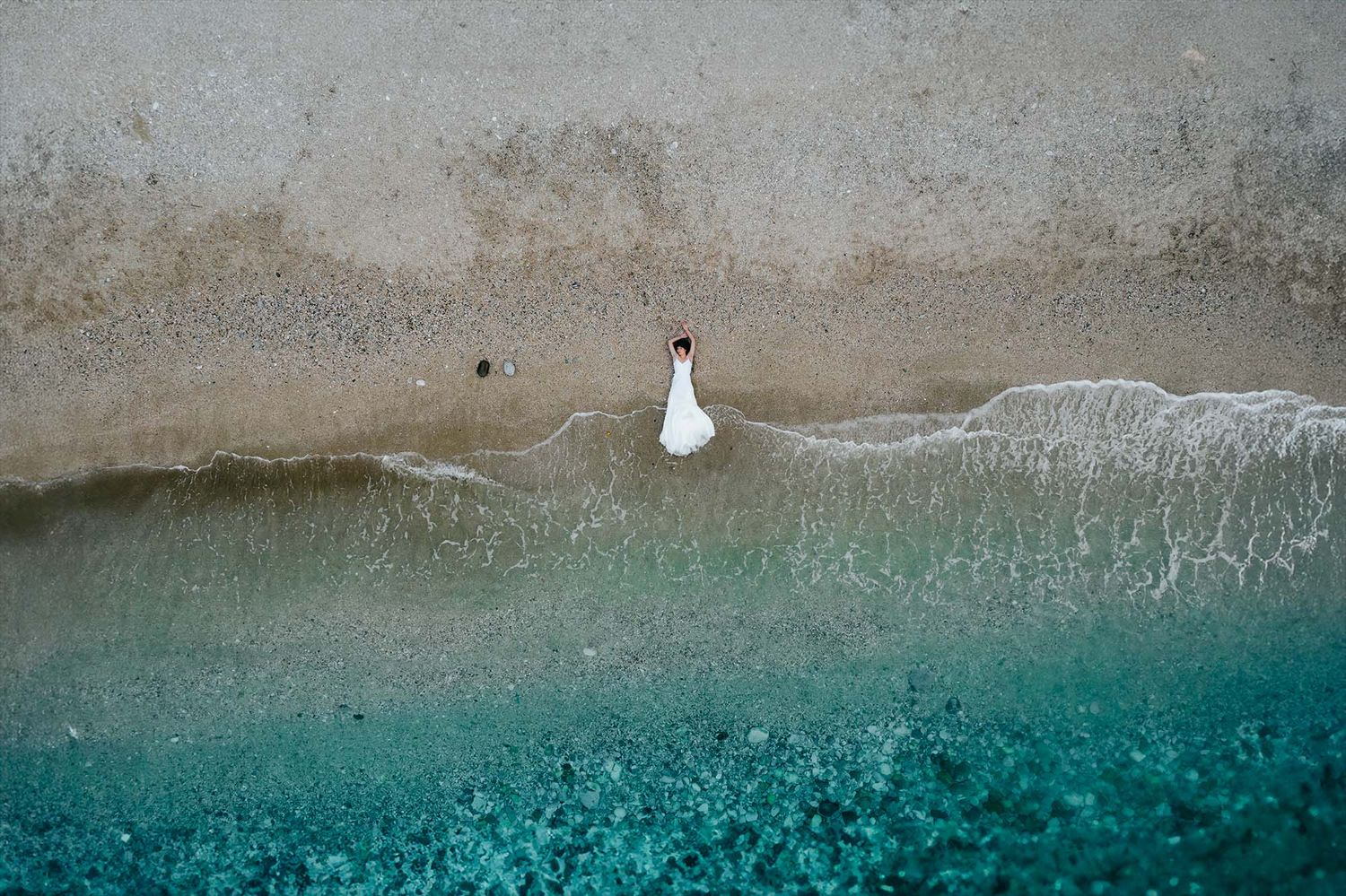 Aerial view of a person in white dress walking along turquoise beach waters and beige sandy shore.
