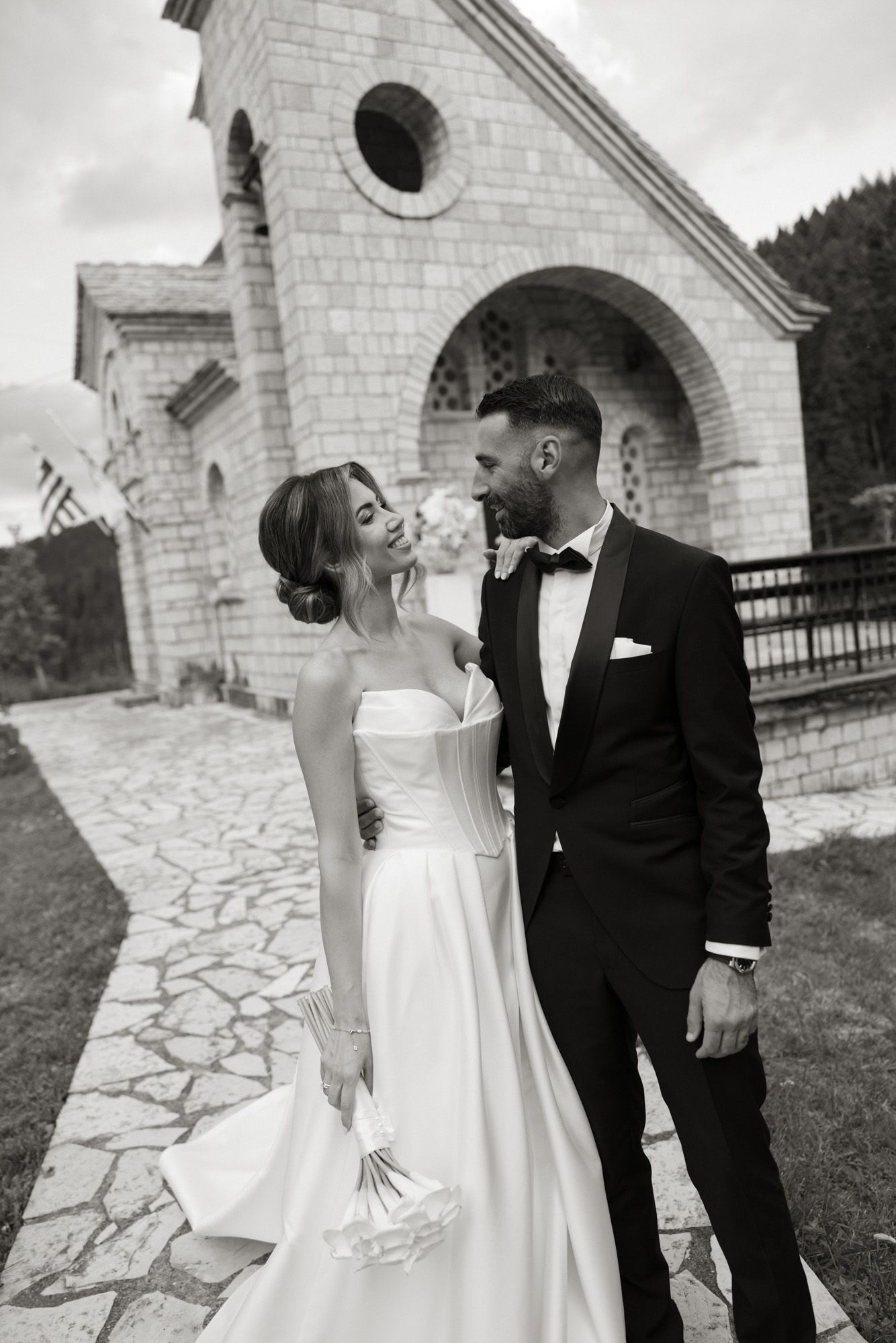 Black and white portrait of an elegant bride and groom embracing outside a stone chapel.