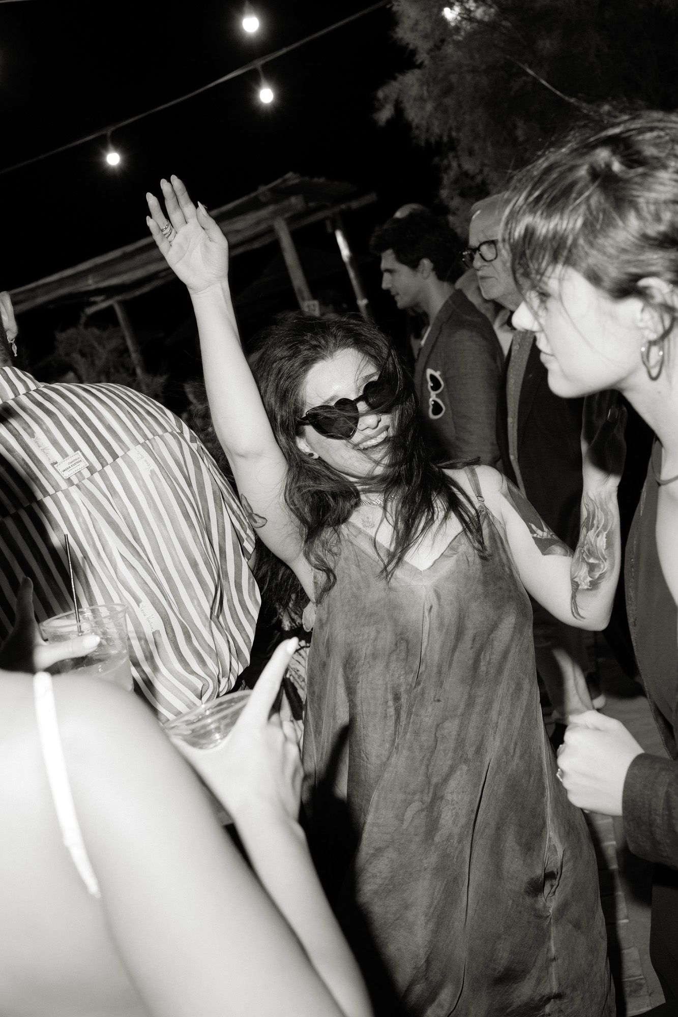 Woman with sunglasses dancing with raised arm at an outdoor nighttime party event.
