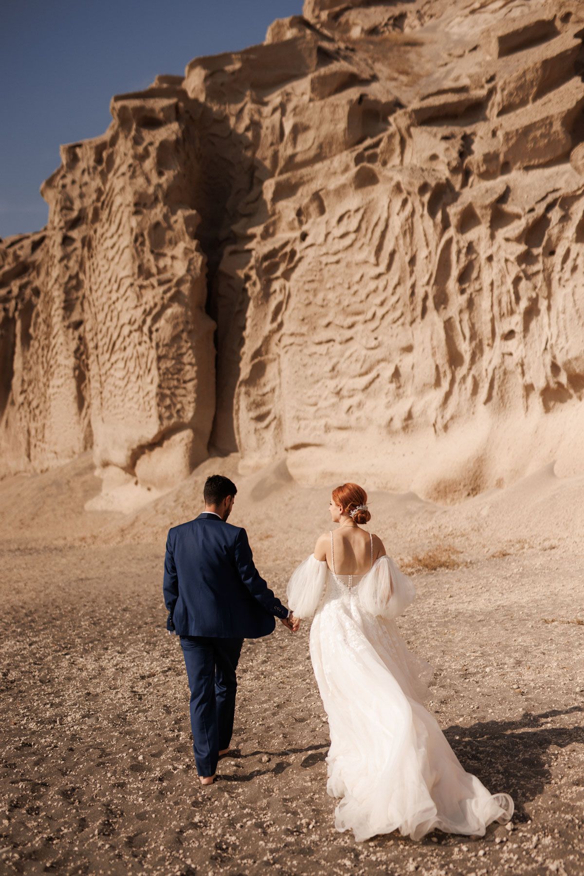 Bride and groom walking hand in hand toward dramatic volcanic rock cliffs in Santorini landscape.