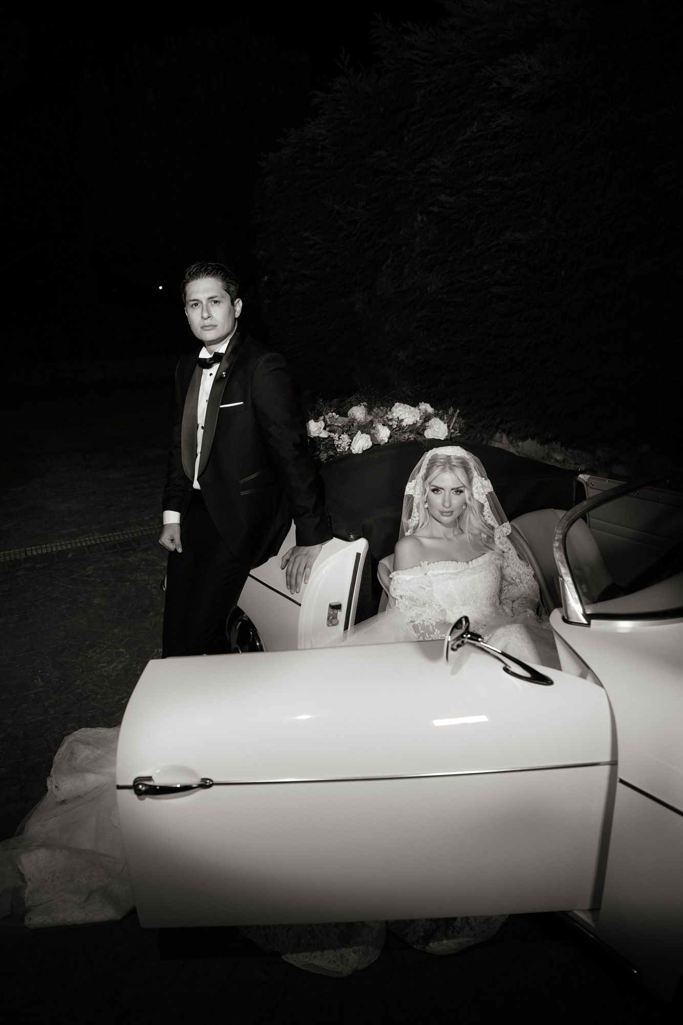 Black and white portrait of newlyweds beside vintage white convertible car at night, bride in gown and groom in tuxedo.