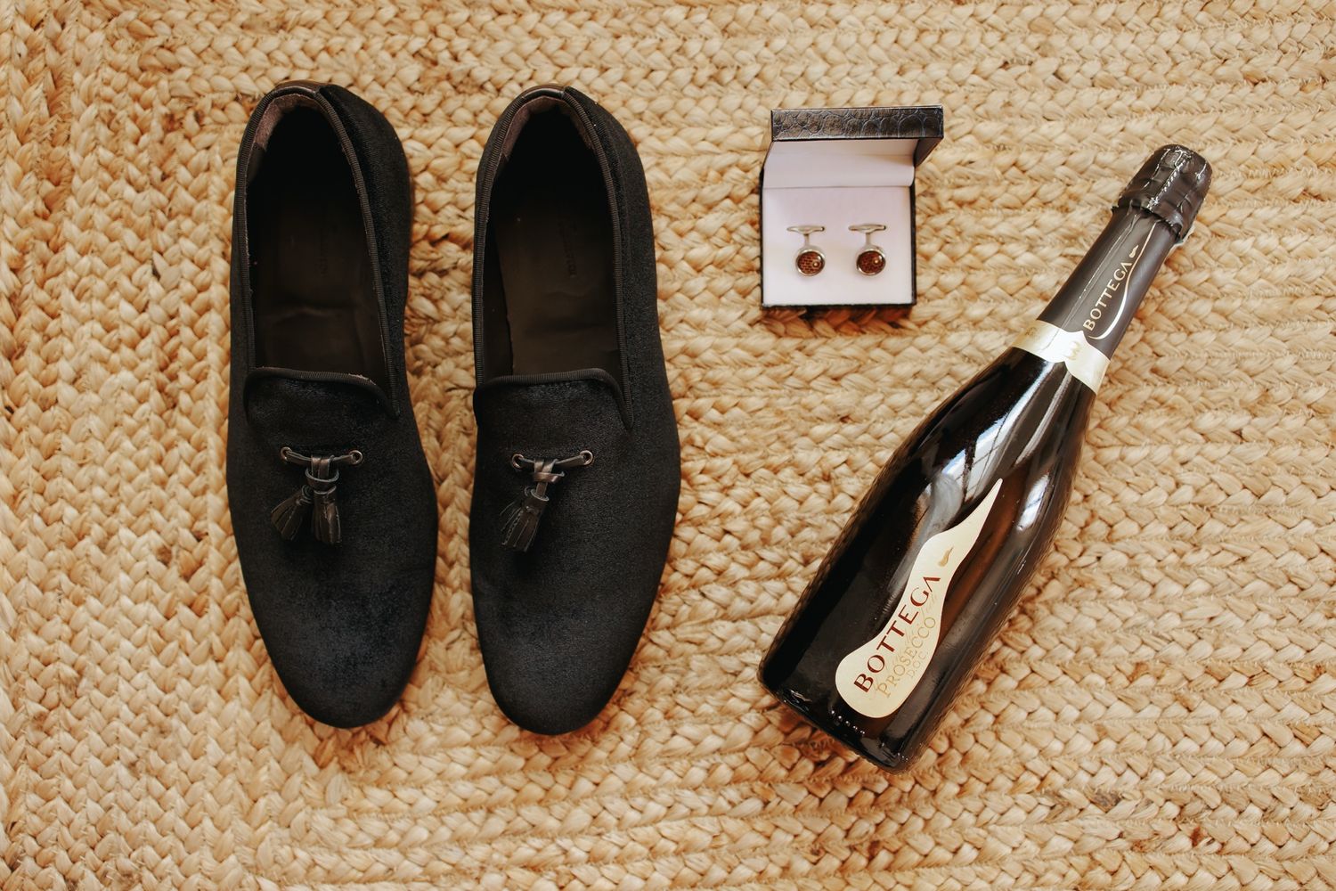 Black tassel loafers, cufflinks, and champagne bottle arranged on woven rug.